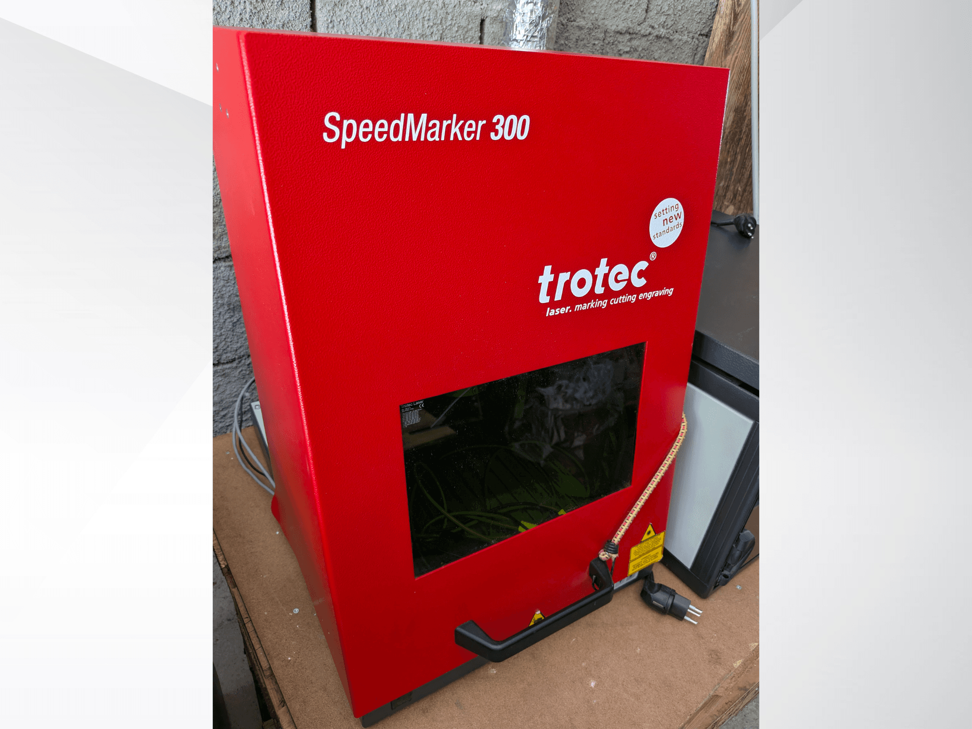 Kırmızı Trotec SpeedMarker 300 lazer markalama makinesinin önden görünümü, büyük bir görüntüleme penceresi ve güç bağlantısı.