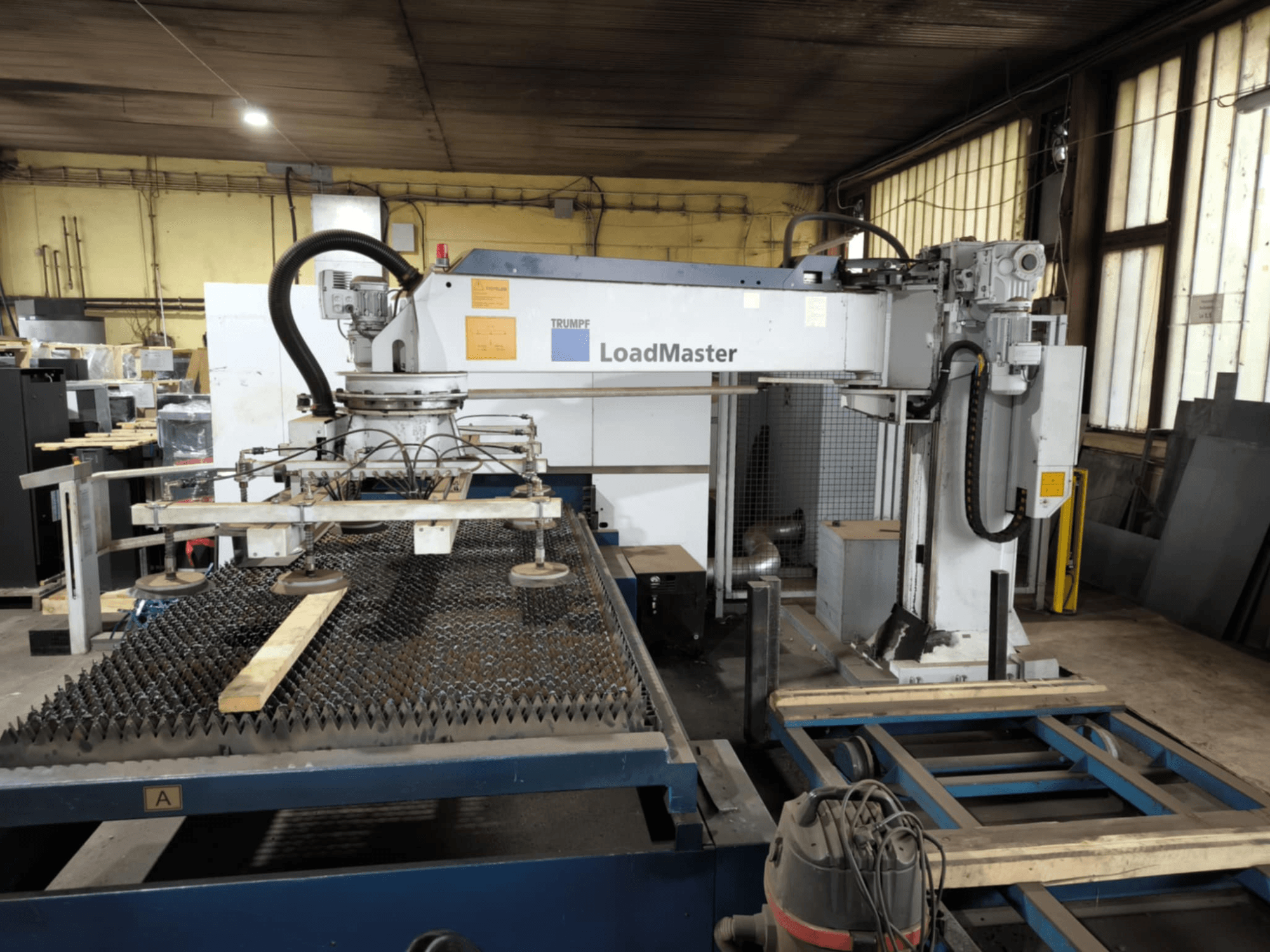 TRUMPF TRULASER 3030 FIBER kesim makinesi, önden görünüm, LoadMaster sistemi sergileniyor ve ızgaralı bir çalışma masası üzerine yerleştirilmiş.