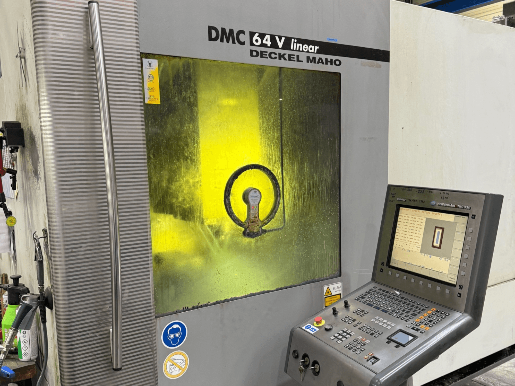 Deckel Maho'dan DMC 64 V linear, kontrol paneli ve iç mekanizmaları gösteren sarı renkli pencere ile makinenin önden görünümü.