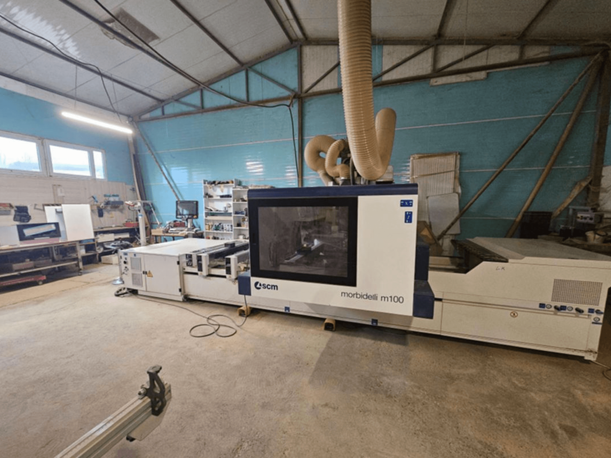 Bir atölyede bulunan SCM MORBIDELLI M 100 CNC işleme merkezi; toz emme sistemi ve çevresindeki çalışma alanı ekipmanlarıyla birlikte önden görünüm.