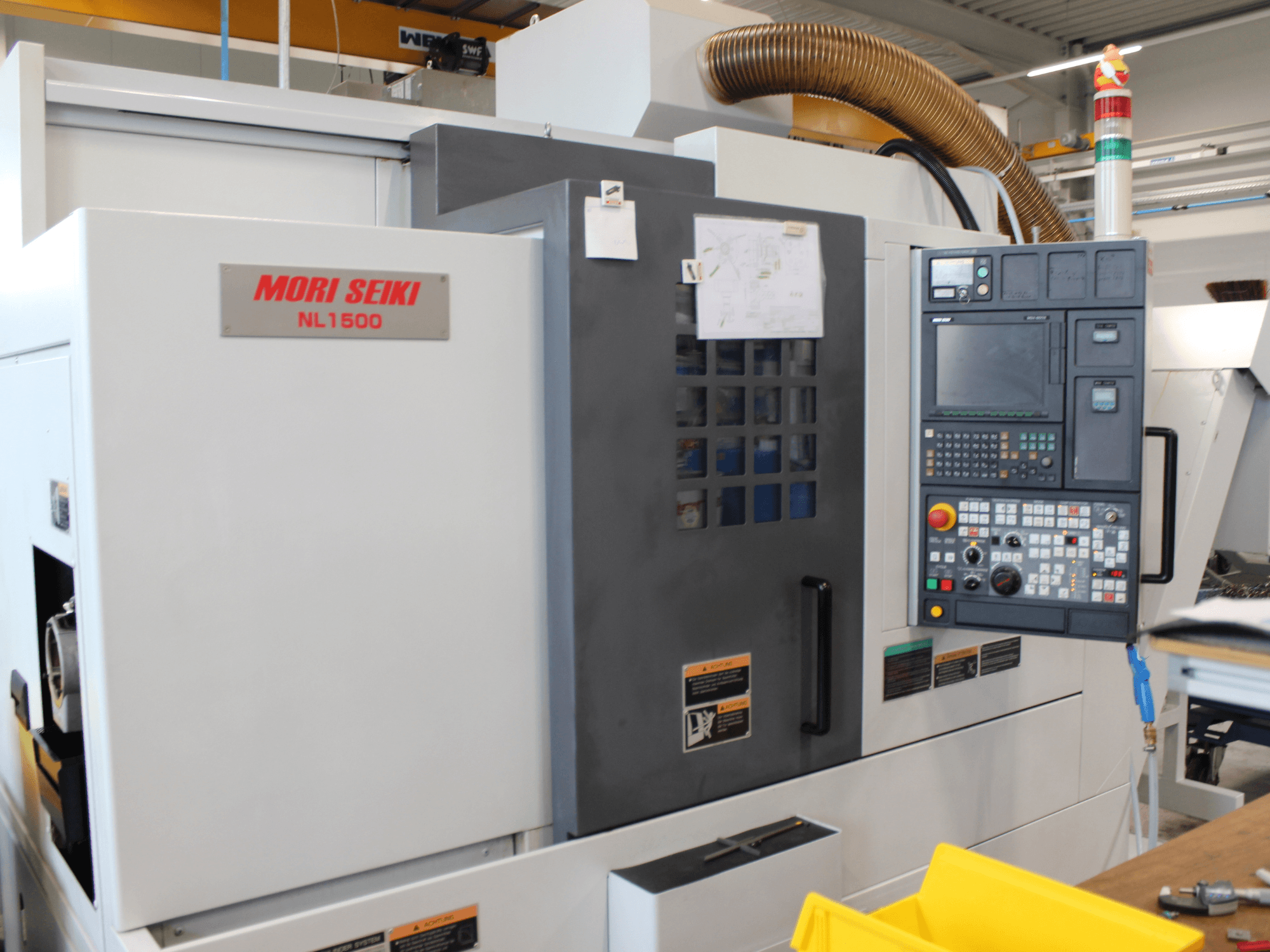MORI SEIKI NL 1500 SY torna-freze merkezinin yandan görünümü; kontrol paneli, iş güvenliği etiketleri ve soğutma sistemi görülmektedir.