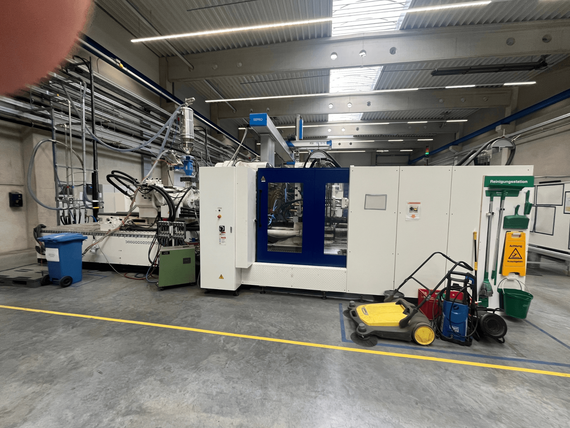 Bir fabrikadaki endüstriyel makine, önden görünüm, mavi ve beyaz paneller, hortumlar ve yakındaki temizlik ekipmanları.