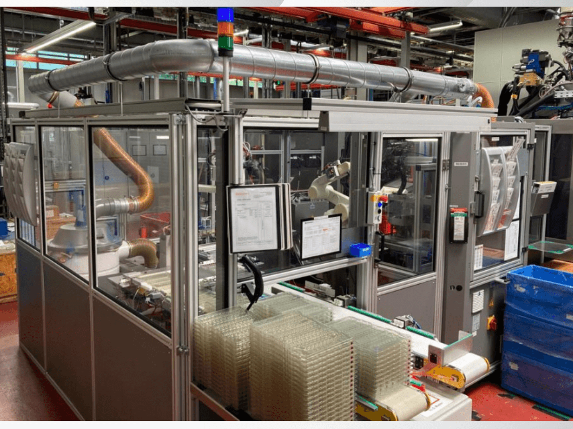 Bir fabrika zemininde metal borular, kontrol paneli ve istiflenmiş şeffaf plastik kaplar içeren otomatik bir makinenin önden görünümü.