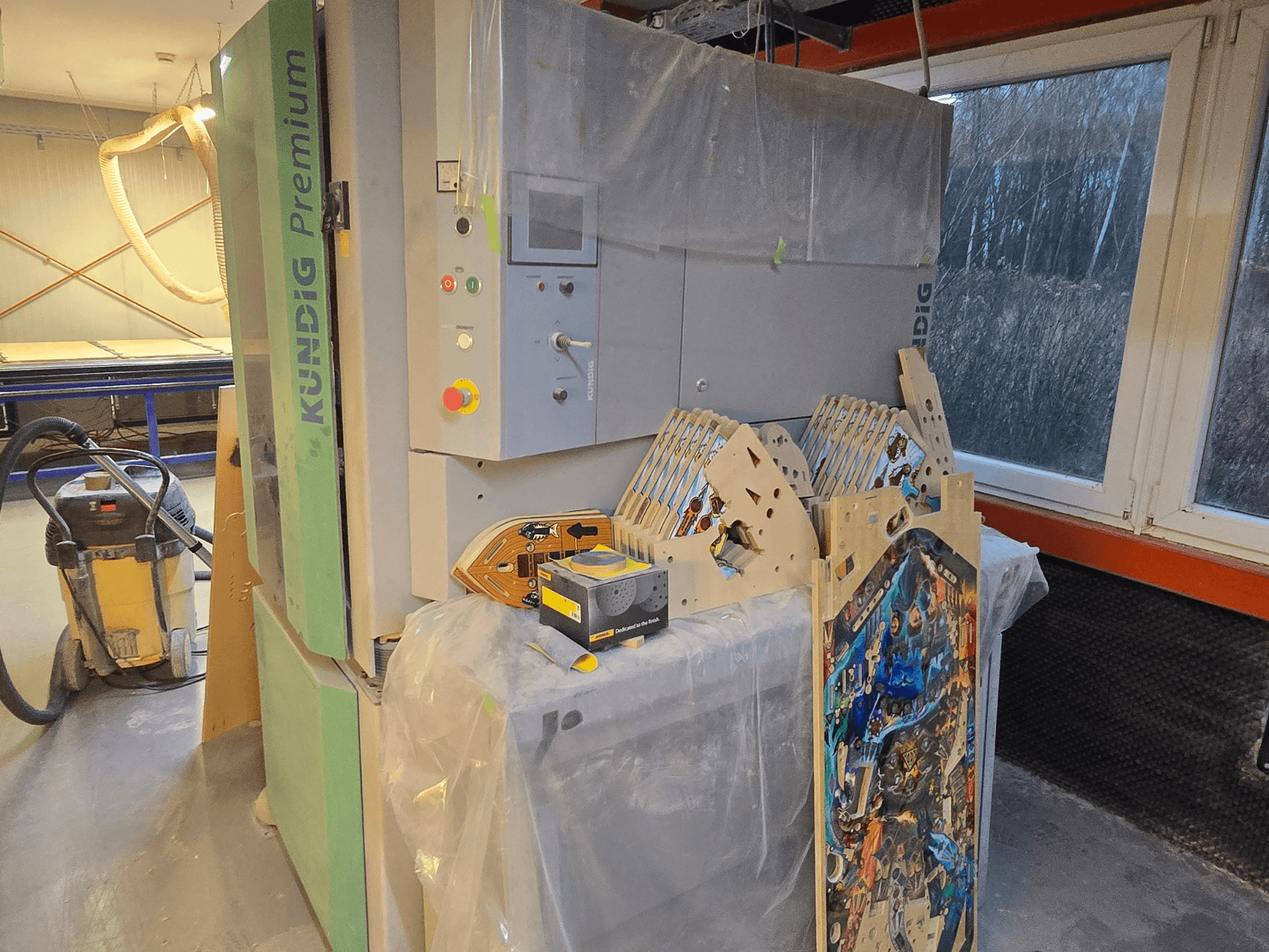 Kundig Premium-1 1100-C bant zımpara atölyede, kontrol paneli ve ahşap parçaları gösteriliyor, yandan görünüm, arka planda toz vakumu ile.