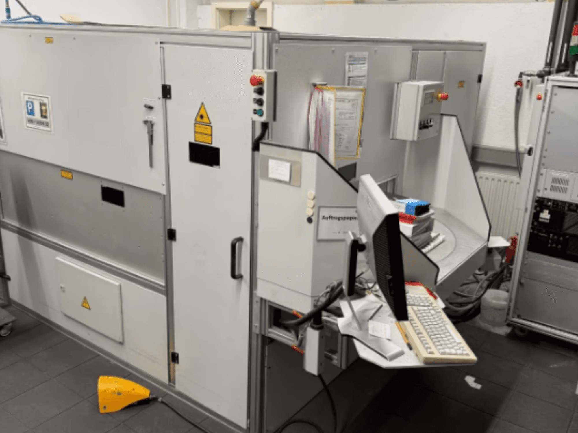 Bilgisayar terminalli endüstriyel makine, yandan görünüm; kontrol düğmeleri ve güvenlik uyarı işaretleri içerir.