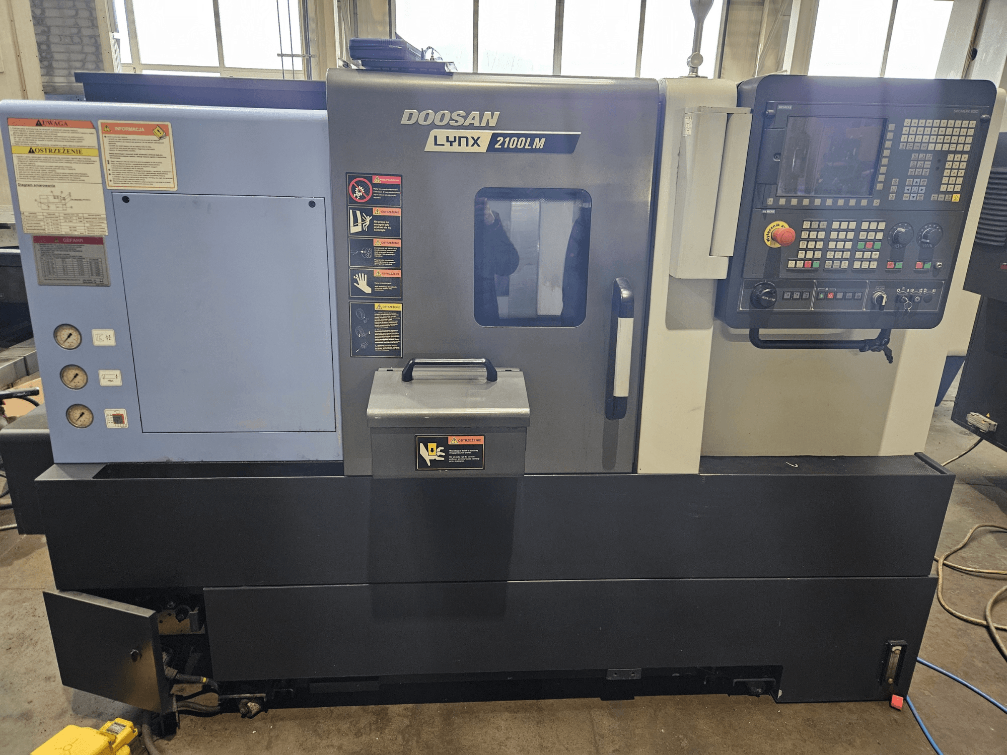Doosan Lynx 2100LM CNC torna tezgahı, önden görünüm, kontrol paneli, hava basıncı göstergeleri ve güvenlik etiketleri görünür.