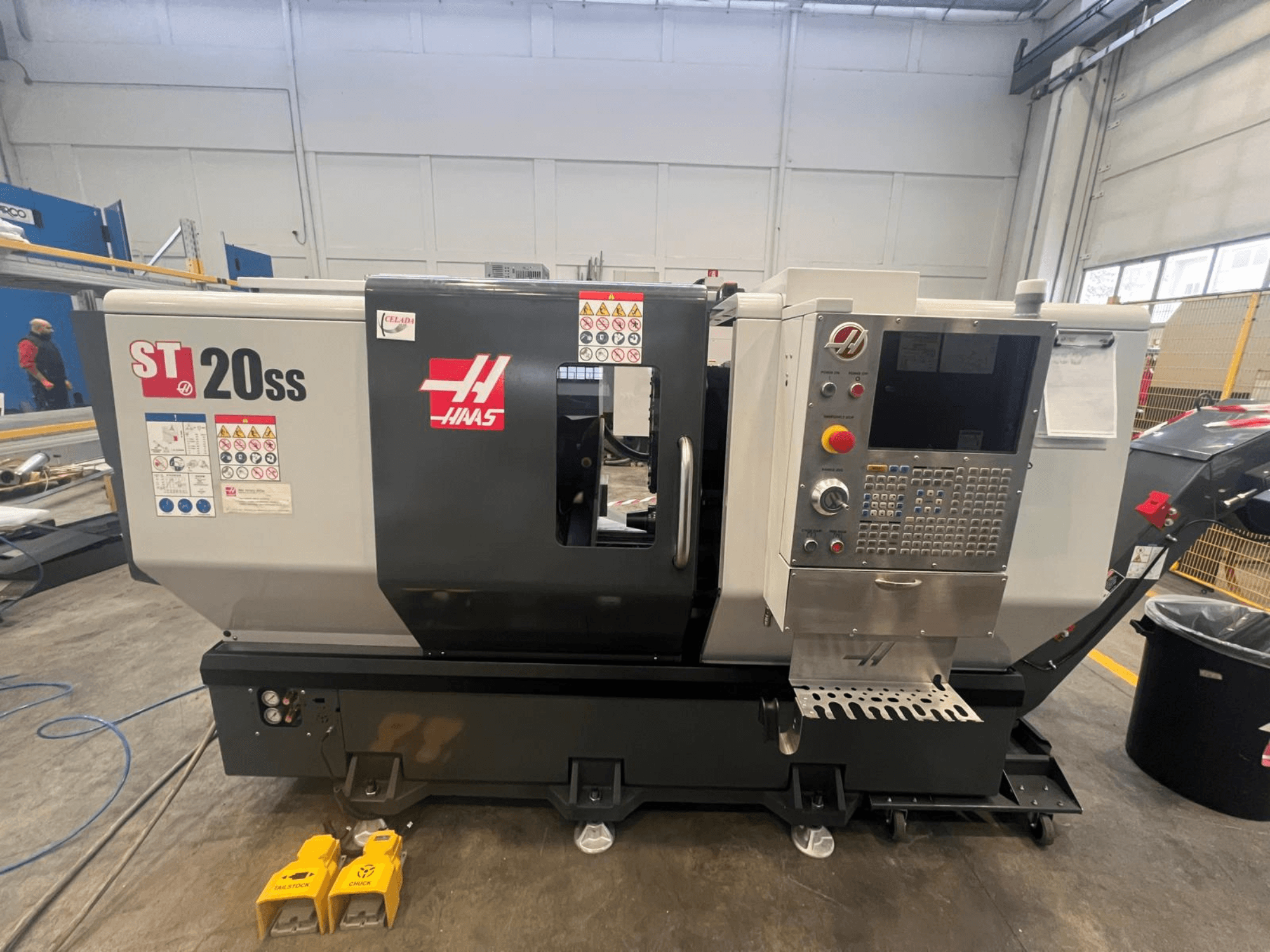 Haas ST 20SS CNC torna tezgahının yandan görünümü, kontrol paneli, güvenlik etiketleri ve zemindeki ayak pedalları.