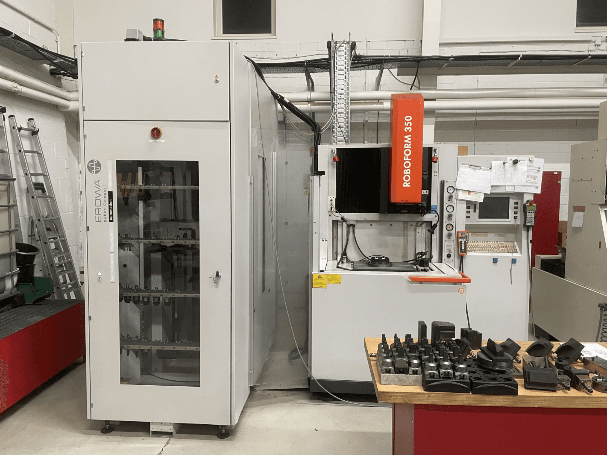 Kalıp oyma EDM makinesi CHARMILLES Roboform 350, önden görünüm, bir masa üzerinde sergilenen aletler ve bitişik ekipman.
