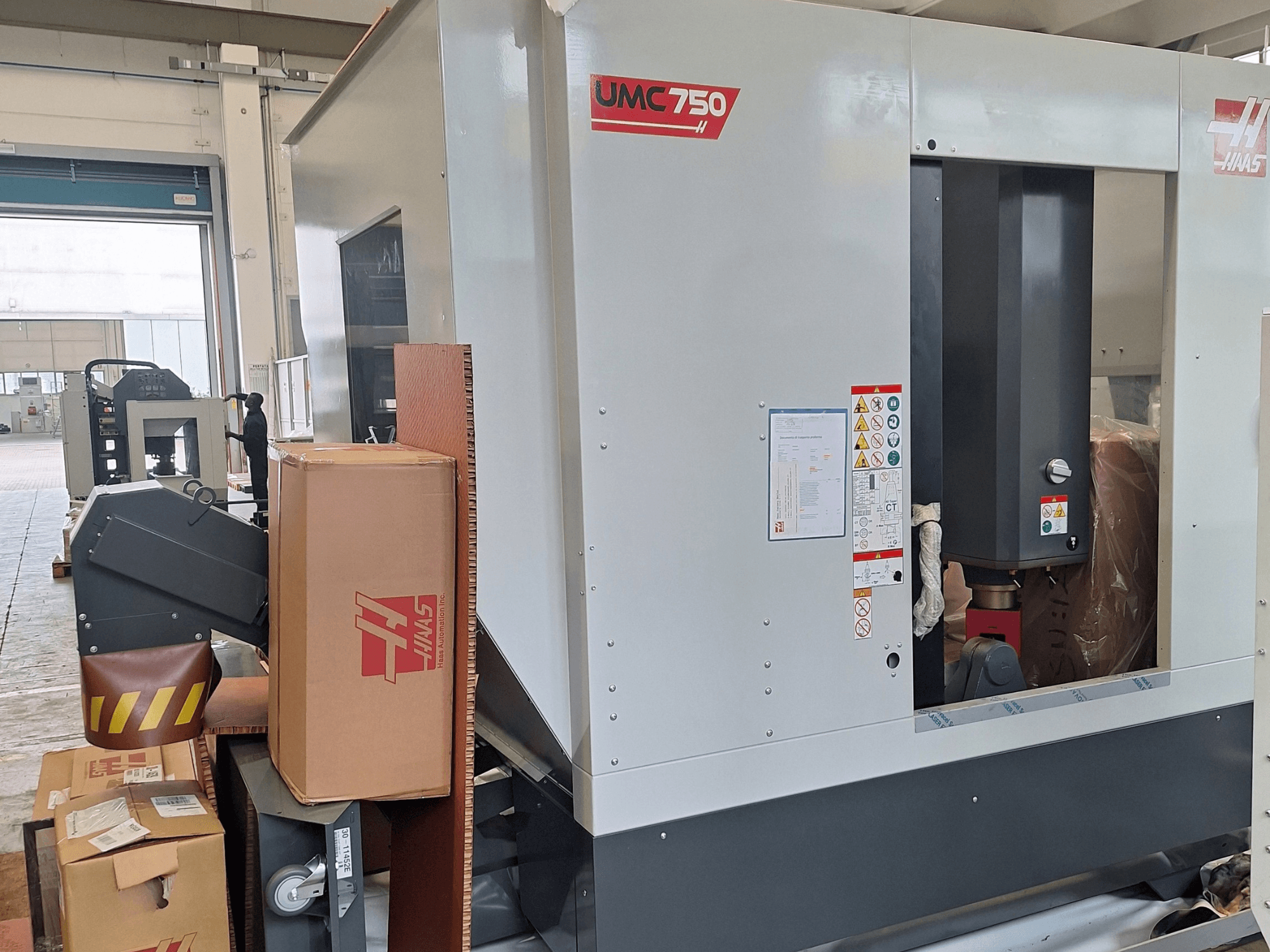 HAAS UMC-750 evrensel işleme merkezi, yandan görünüm; kontrol paneli ve uyarı etiketleri ile birlikte takım saklama bölmesi ve ambalajı gösterilmektedir.
