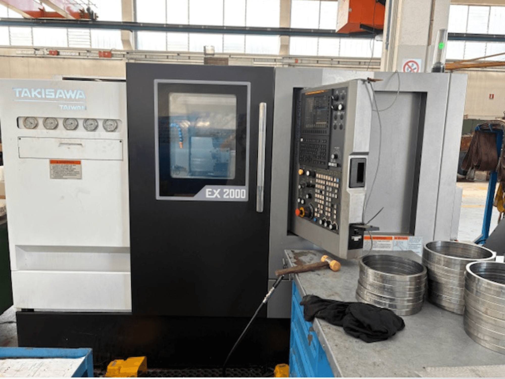 Takisawa EX 2000 CNC makinesi, önden görünüm, kontrol paneli, göstergeler ve atölye masası üzerindeki metal parçalar.