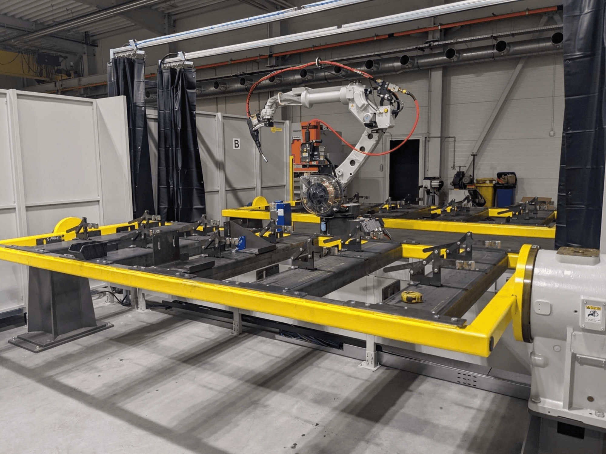 Montaj için metal parçaların bulunduğu sarı bir çalışma masasının üzerine yerleştirilmiş PANASONIC TAWERS TM-2000WG3 robot kolunun önden görünümü.