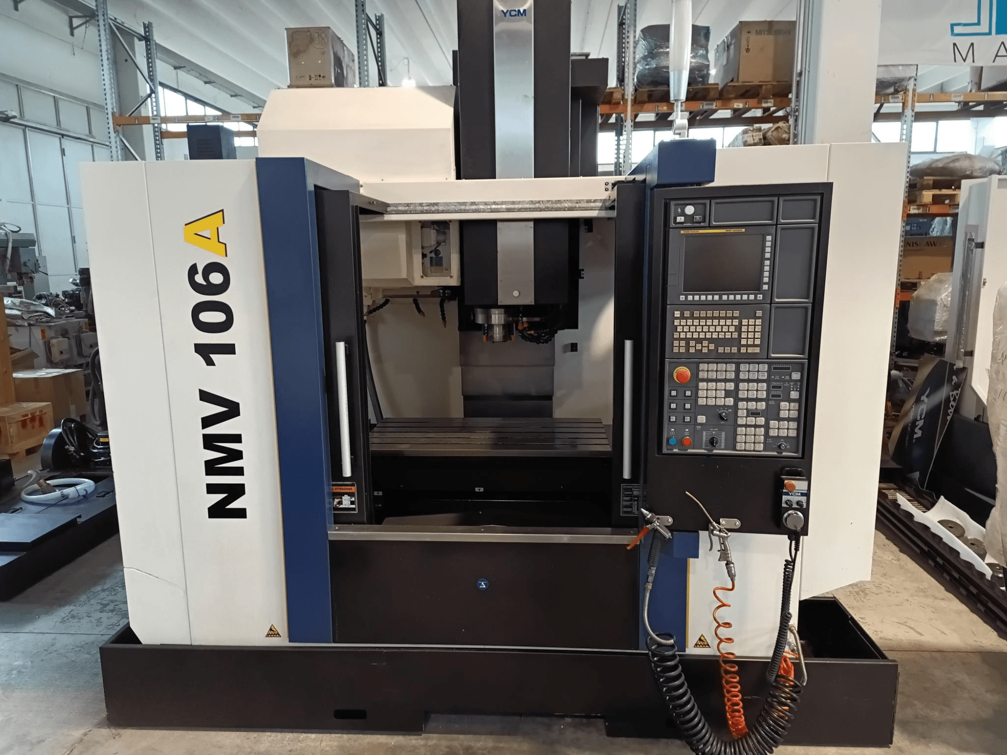 YCM NMV 106A işleme merkezi, önden görünüm, düğmeli kontrol paneli ve soğutma sıvısı hortumları.