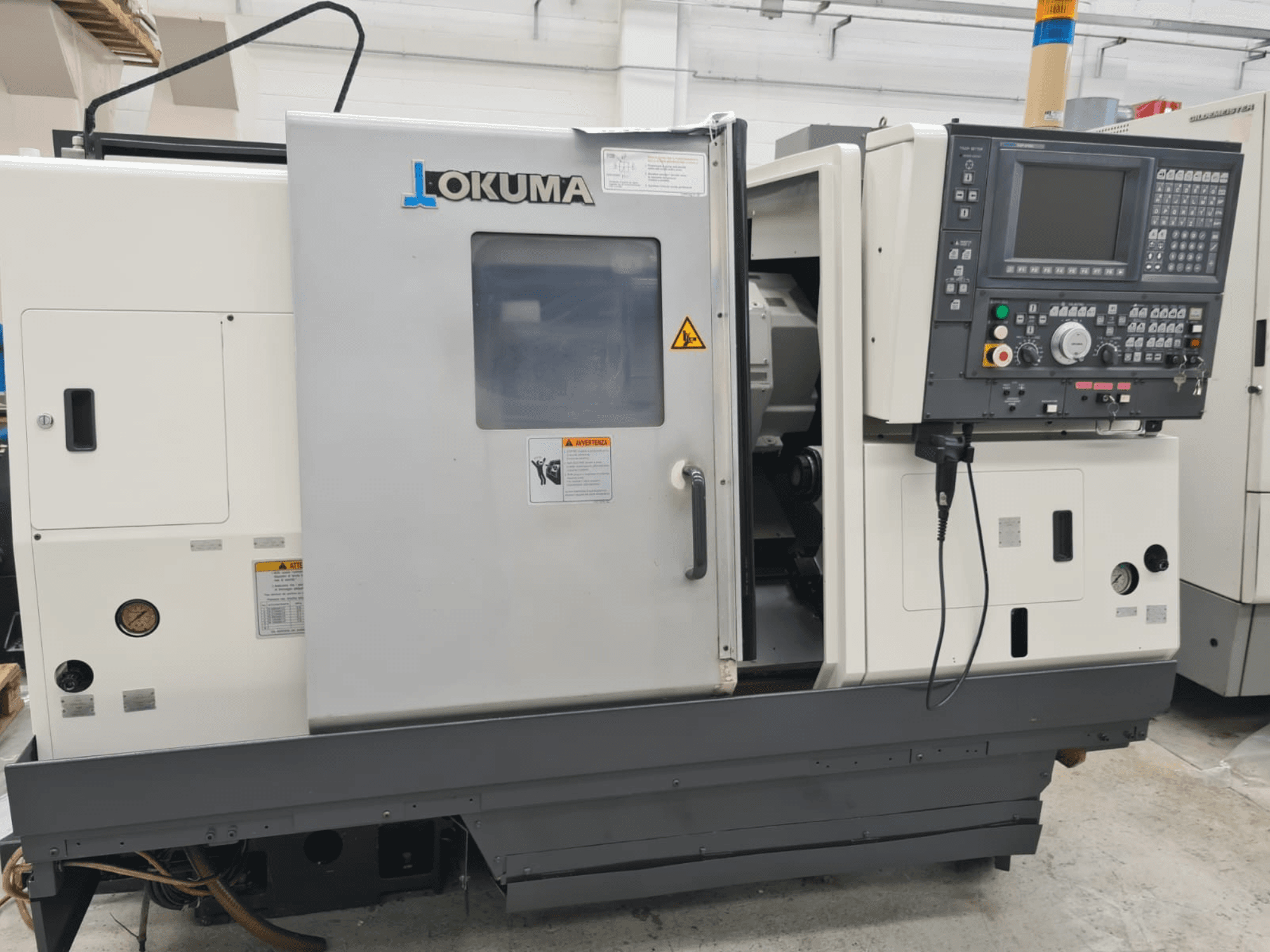 Okuma LB 300M X 500 yatay torna makinesinin yandan görünümü, kontrol paneli, iş mili erişimi ve güvenlik uyarıları.