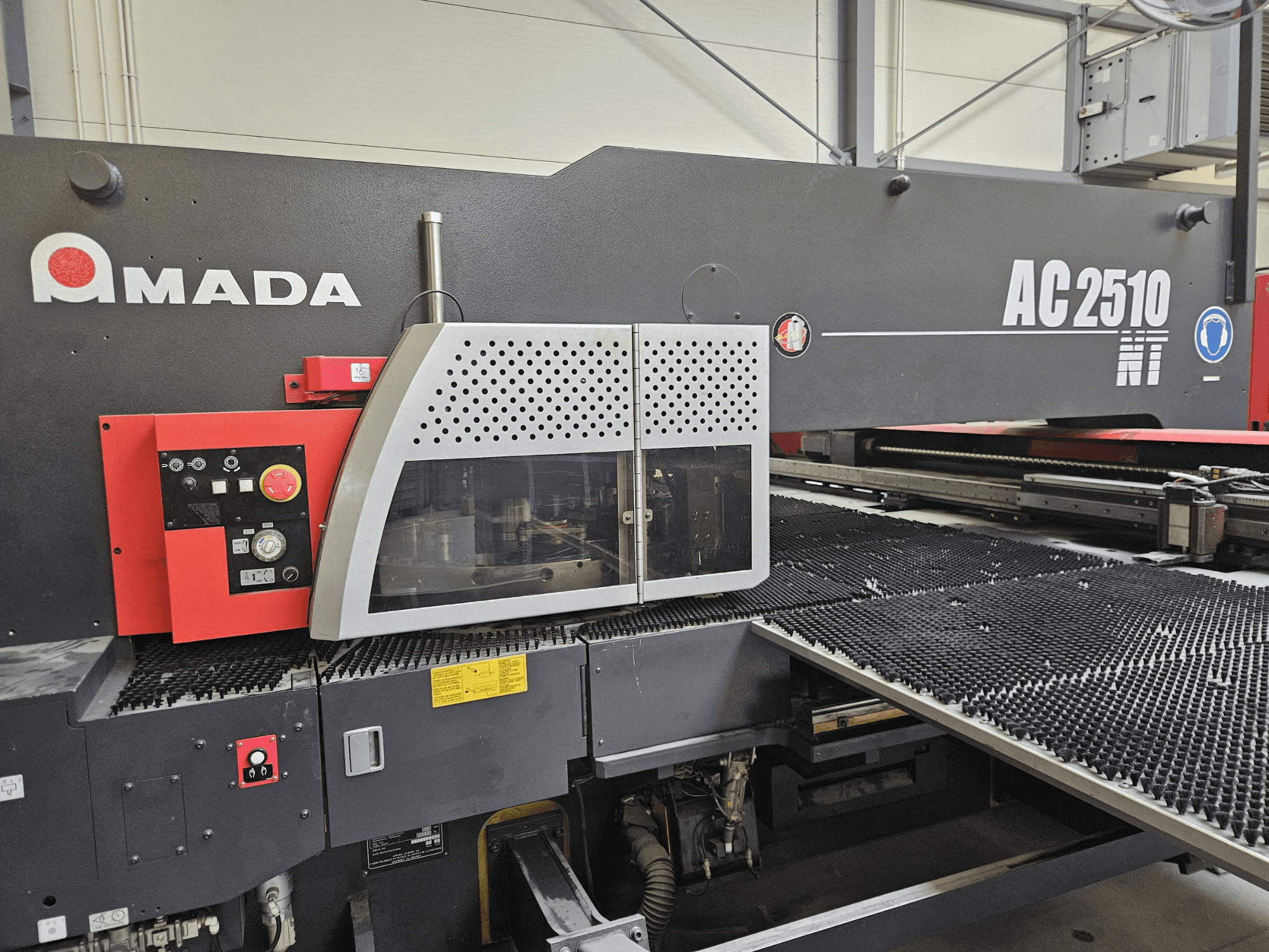 Amada AC2510 NT lazer kesim makinesi, önden görünüm, düğmeler, butonlar ve güvenlik kapağı içeren kontrol paneli.