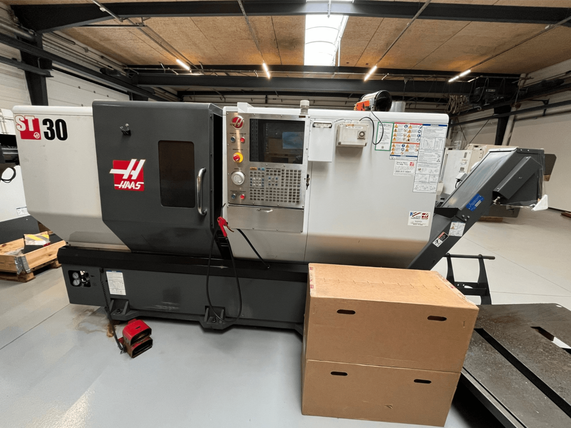 Haas ST-30 CNC torna tezgahı, önden görünüm, düğmeli kontrol paneli, acil durdurma ve istiflenmiş iki karton kutu.