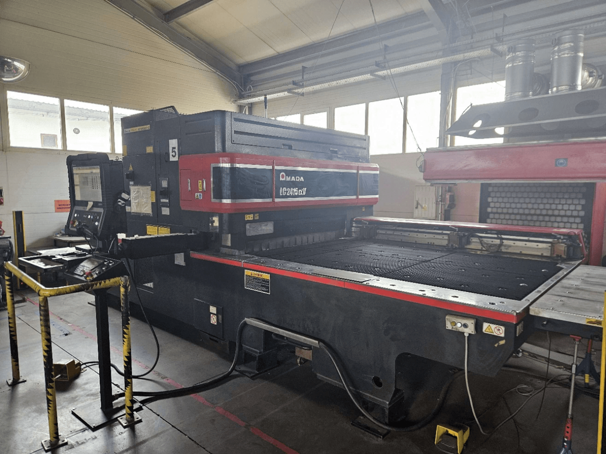 AMADA LC2415 Alpha 5 CO₂ lazer kesim makinesi; kontrol paneli ve güvenlik bariyerleriyle donatılmış kesim yatağını gösteren önden görünüm.