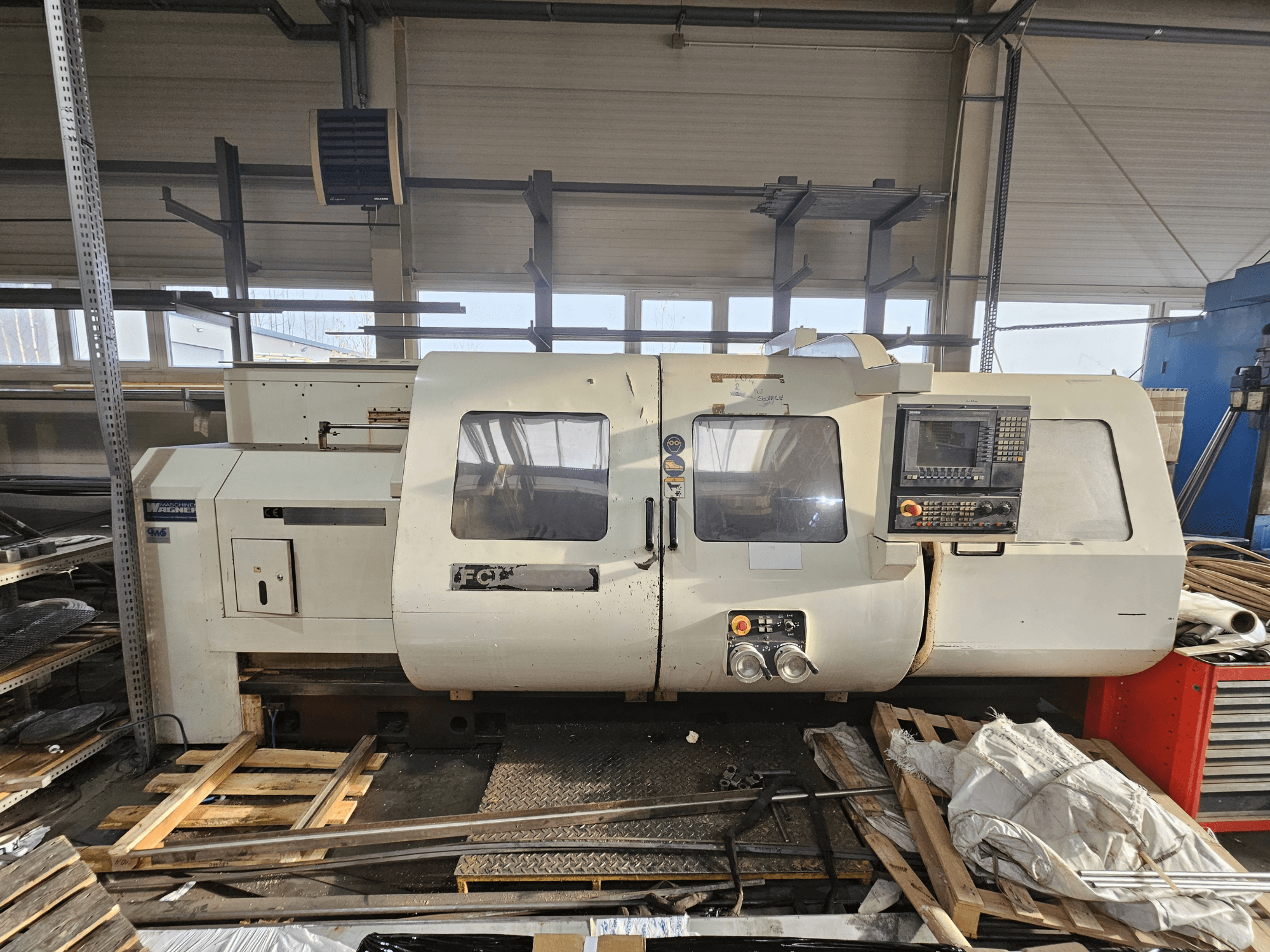 Wabeco CNC torna tezgahı, yandan görünüm, kontrol paneli, pencereler ve çalışma alanı; atölye ortamında ahşap paletler üzerindeki makineler.