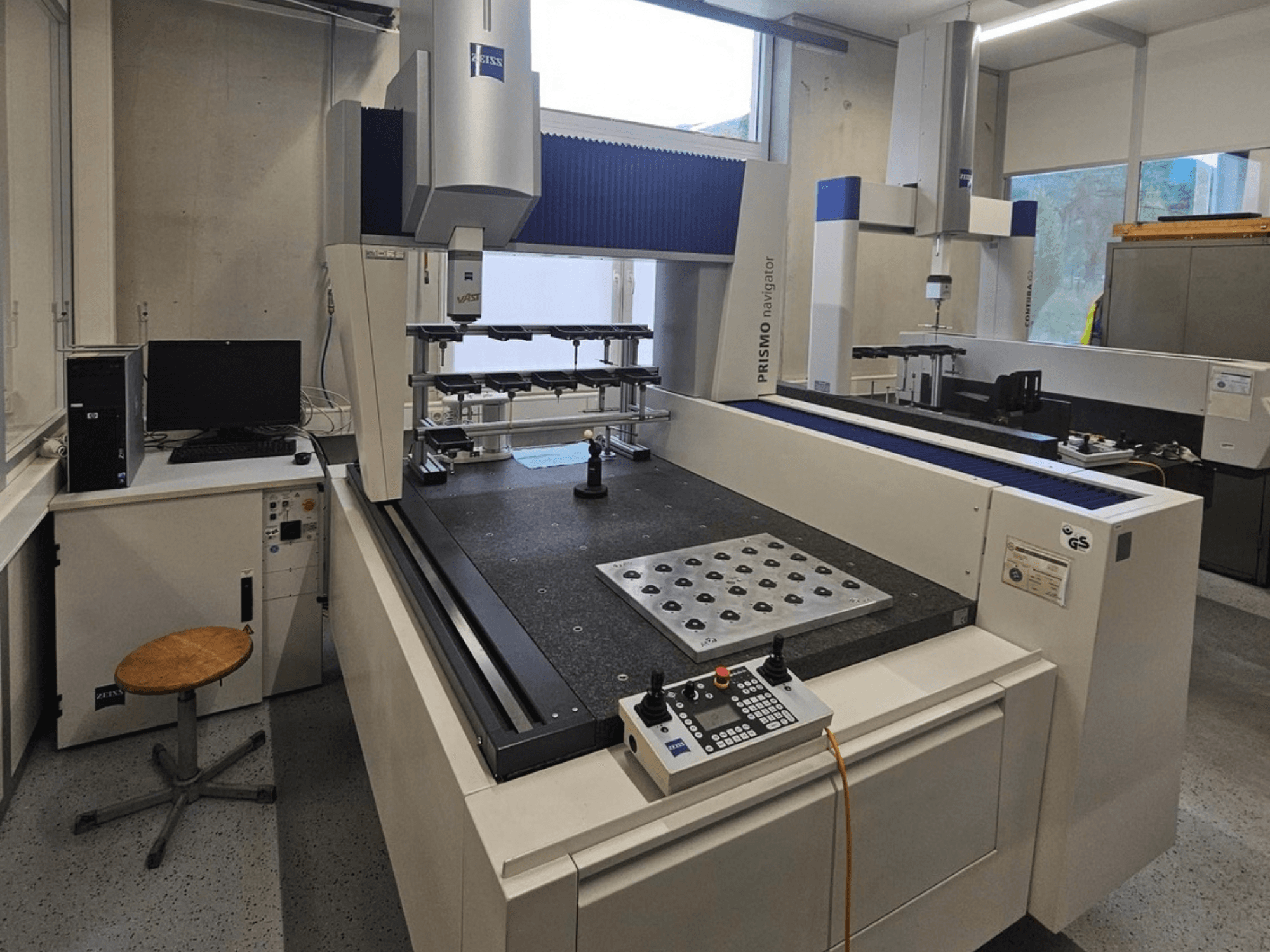 Bir laboratuvarda bulunan ZEISS Prismo Navigator 7 CMM; kontrol paneli, çalışma masası ve monitörü gösteren önden görünüm.