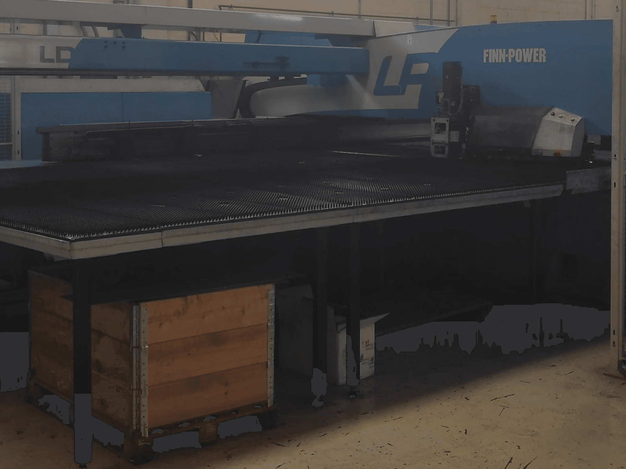 Bir fabrikada Finn-Power LP kesme makinesi, dokulu metal masa ve altındaki ahşap sandık ile önden görünüm.