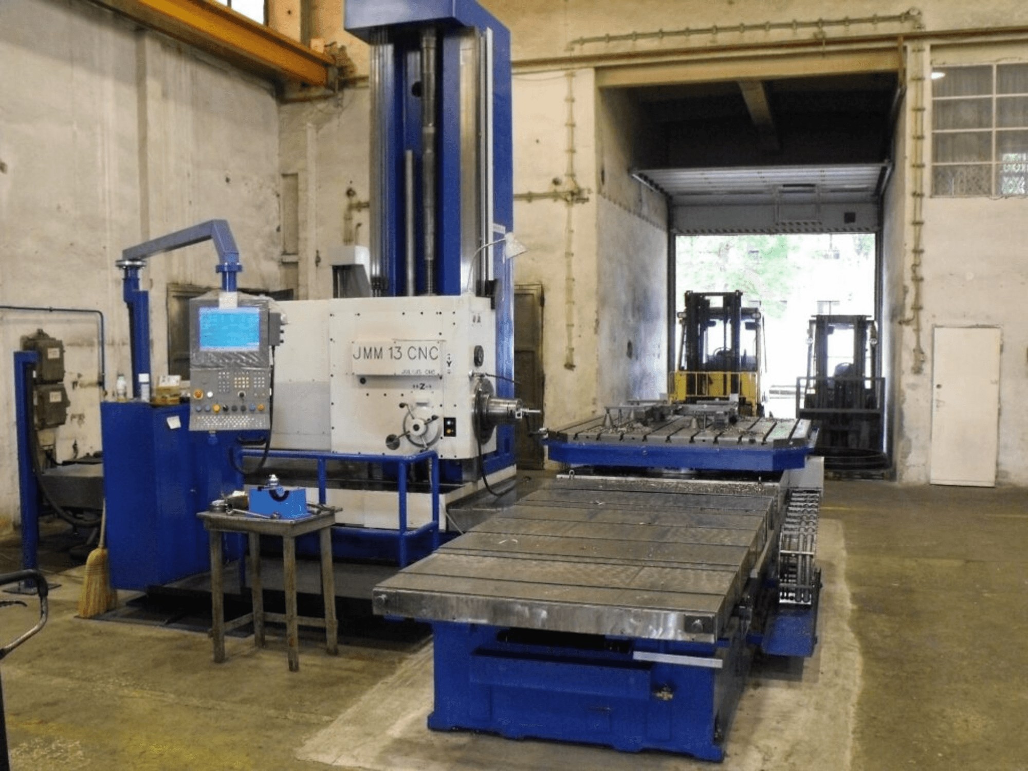 Bir atölyedeki JMM 13 CNC freze makinesi, önden görünüm, bir kontrol paneli, büyük çalışma masası ve endüstriyel arka plana sahiptir.