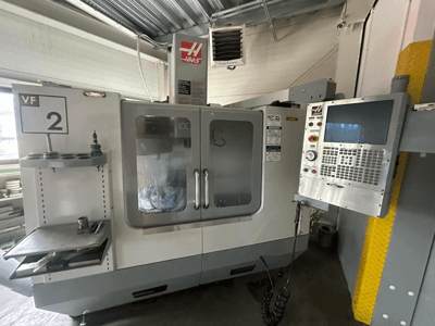 Haas VF-2 CNC makinesi, önden görünüm, kontroller, takım tutucu ve kabinin içinde görünür takımlar bulunan şeffaf kapılar.