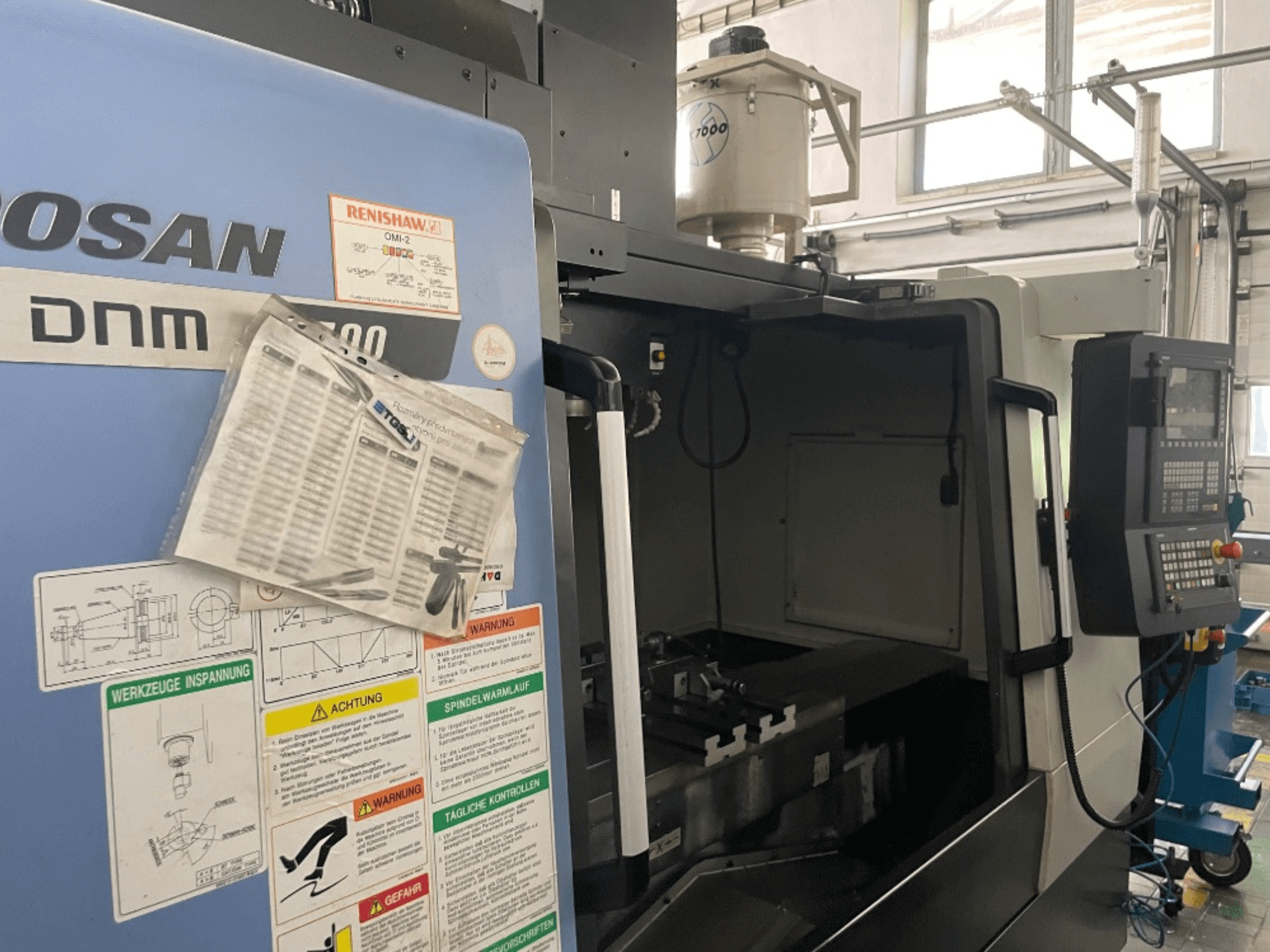 DOOSAN DNM 5700 dik işleme merkezi, önden görünüm, kontrol panelini, güvenlik etiketlerini ve makine detaylarını gösterir.