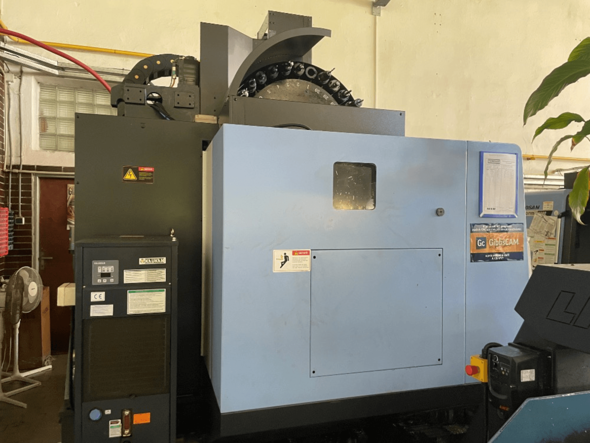 DOOSAN DNM 5700 dik işleme merkezinin önden görünümü, kontrol paneli, güvenlik etiketleri ve makine özellikleri.