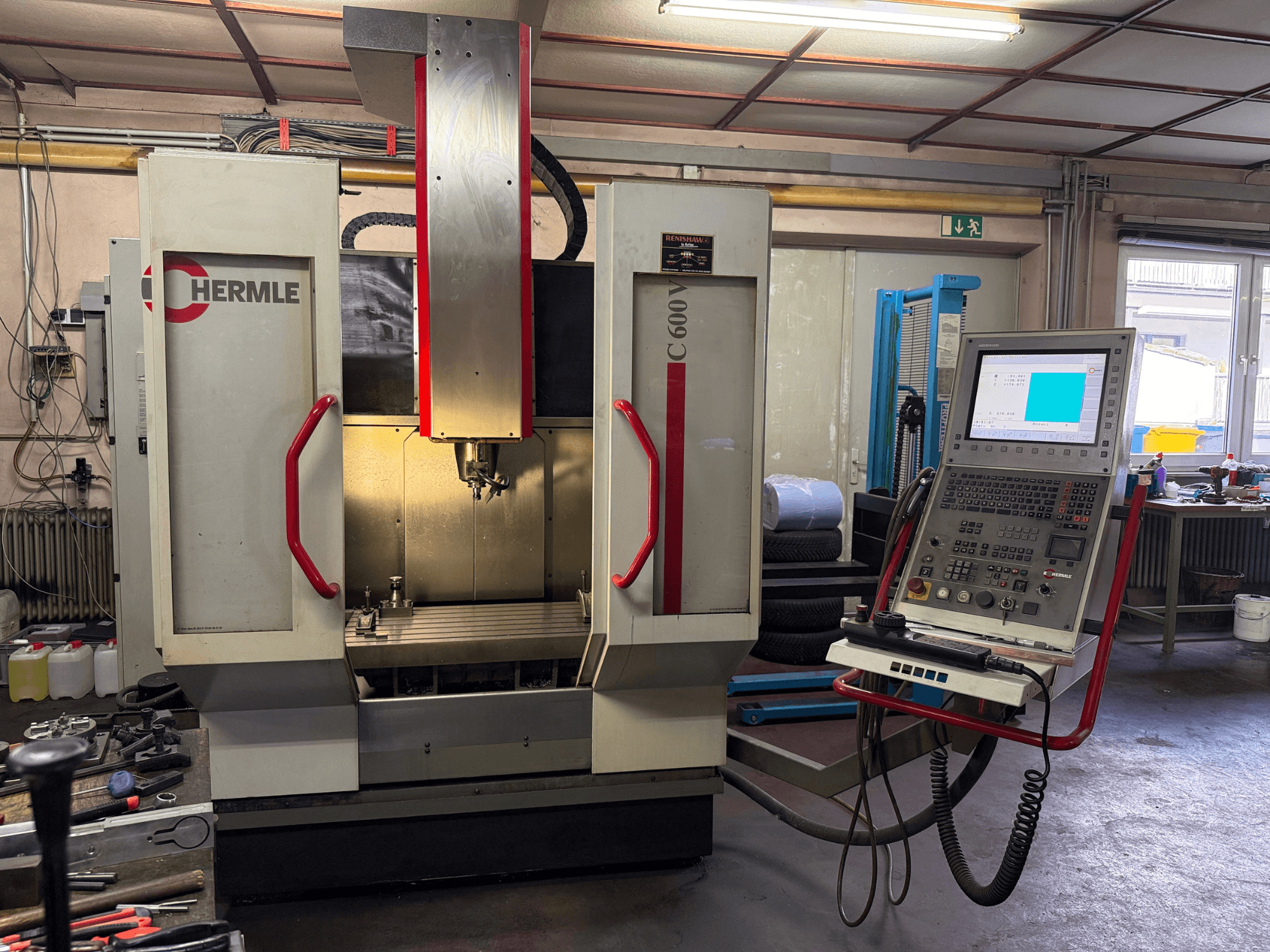 Hermle C 600 CNC freze makinesinin önden görünümü, çok sayıda düğmeye sahip bir kontrol paneli ve geniş bir çalışma alanına sahiptir.