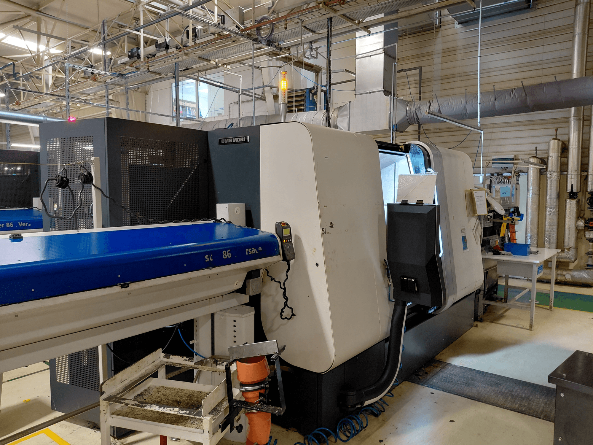 DMG MORI Sprint 65 lineer çok milli torna makinesi, atölye ortamında kontrol paneli ve konveyörün göründüğü yandan görünüm.