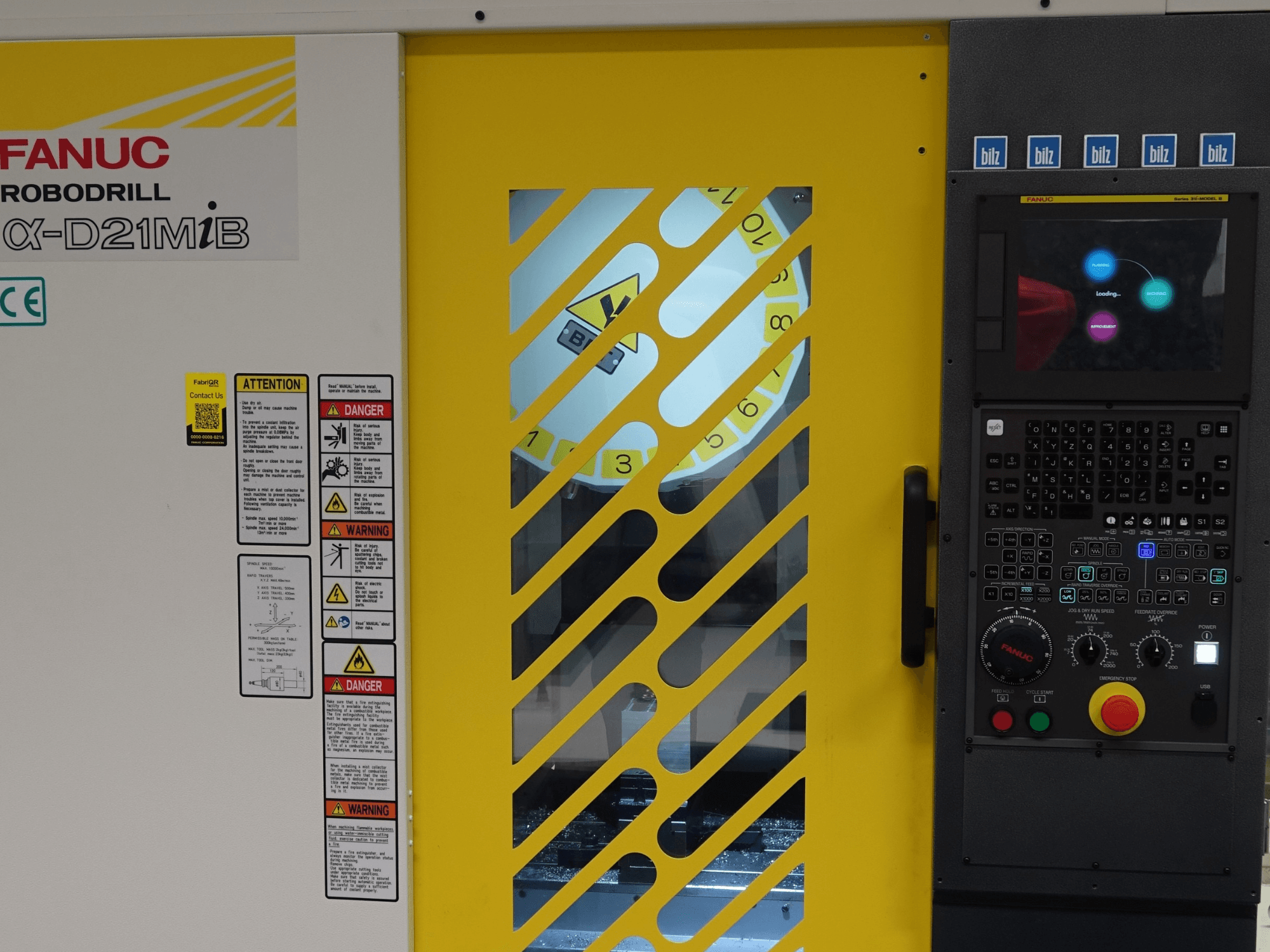 FANUC α-D21MiB Dikey İşleme Merkezi’nin önden görünümü; güvenlik uyarıları, kontrol paneli ve sarı renkli güvenlik kapısı görülmektedir.