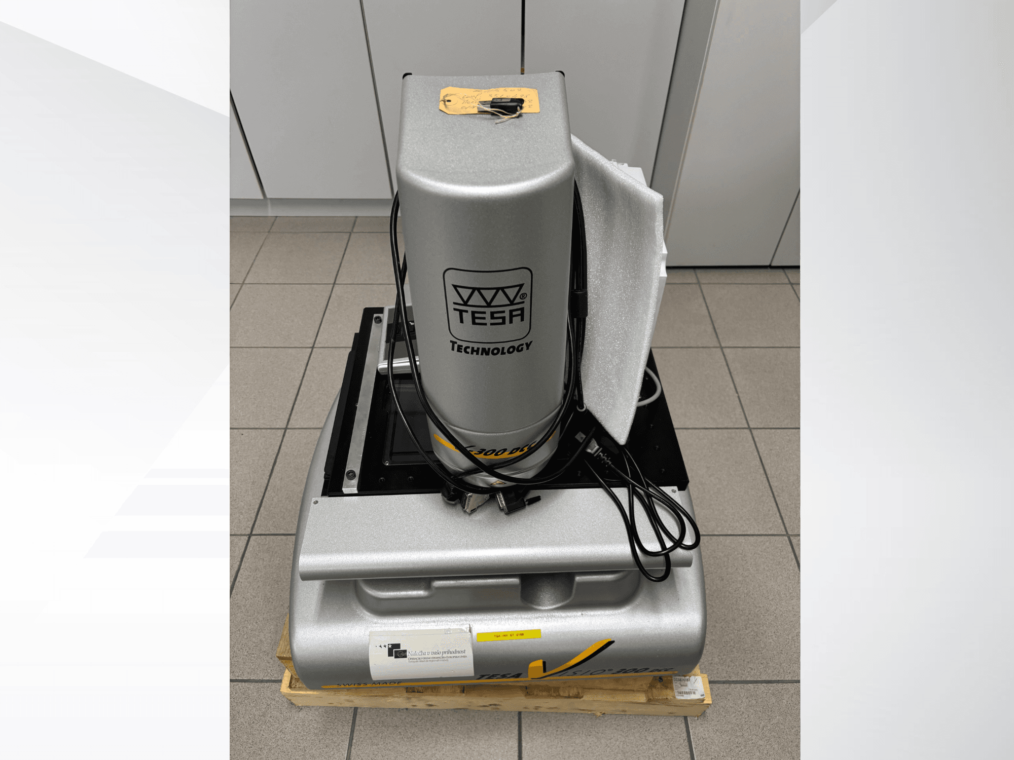 TESA Visio 300 DCC optik ölçüm makinesinin kablolarla birlikte önden görünümü, ahşap bir palet üzerinde ve koruyucu ambalaj görünür durumda.