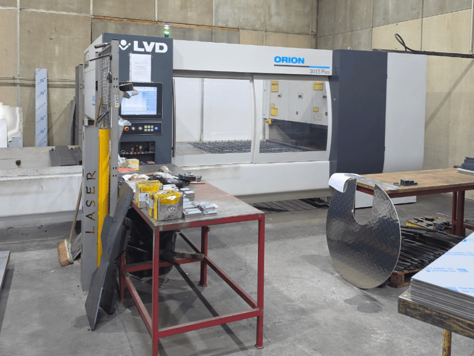 LVD Orion 3015 Plus CO₂ lazer kesim makinesi, kontrol paneli ile birlikte önden görünüm; çalışma masası üzerinde metal parçalarla çevrili.