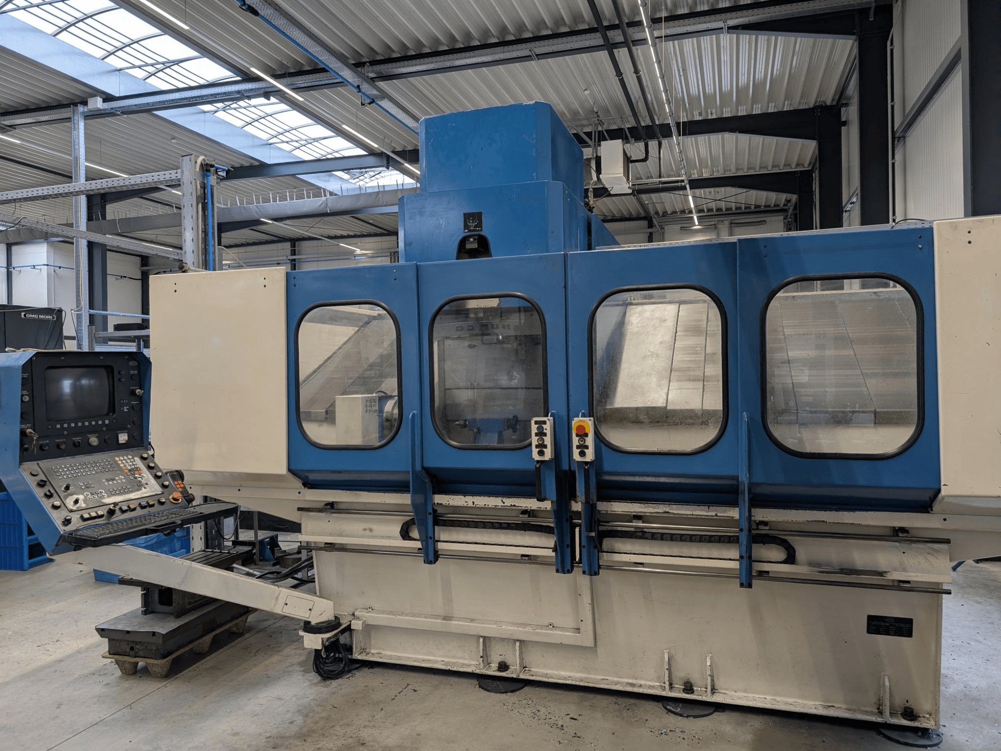 Kontrol panelli mavi endüstriyel CNC makinesi, önden görünüm. Büyük cam pencereler, görünür makine bileşenleri ve düğmeler.