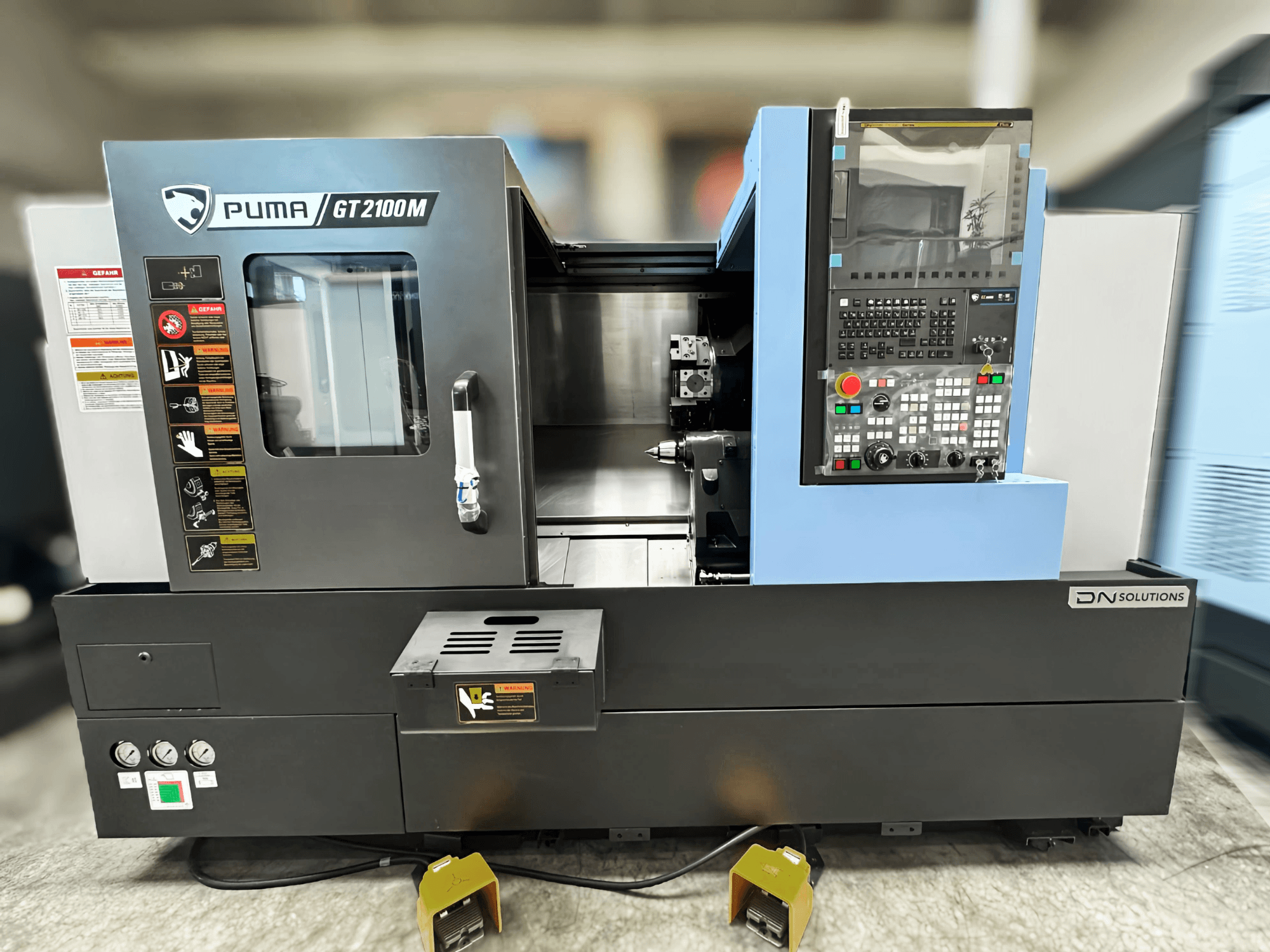 Torna freze merkezi DN SOLUTIONS PUMA GT2100M, kontrol paneli, işleme odası ve güvenlik etiketlerini gösteren önden görünüm.