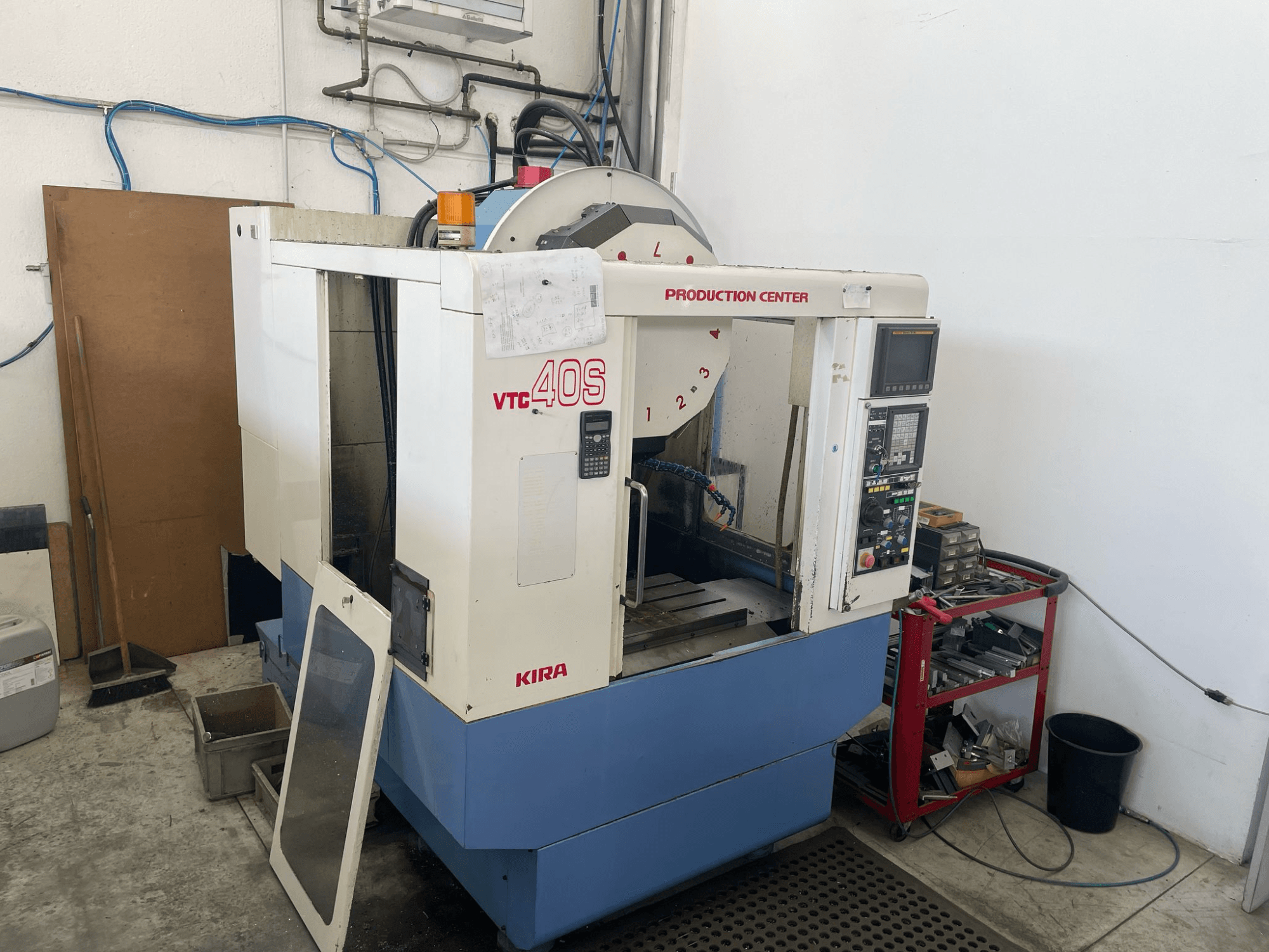 KIRA VTC 40S işleme merkezi yandan görünümde, kontrol paneli, açık kapı ve yakındaki bir depolama arabasındaki takımlar.