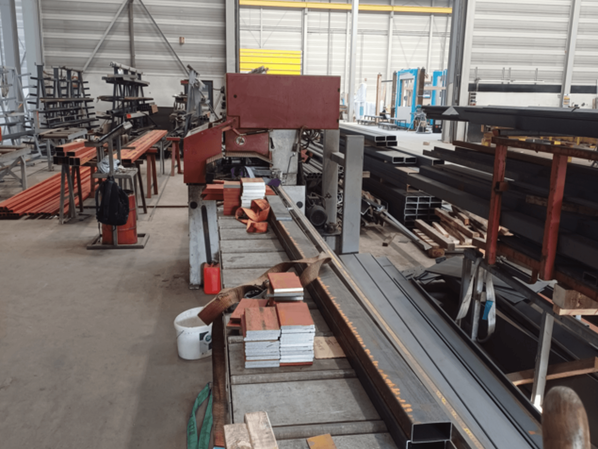 Ön planda kırmızı bir testere makinesi ve istiflenmiş malzemeler ile çeşitli metal profillerin yer aldığı imalat atölyesi.