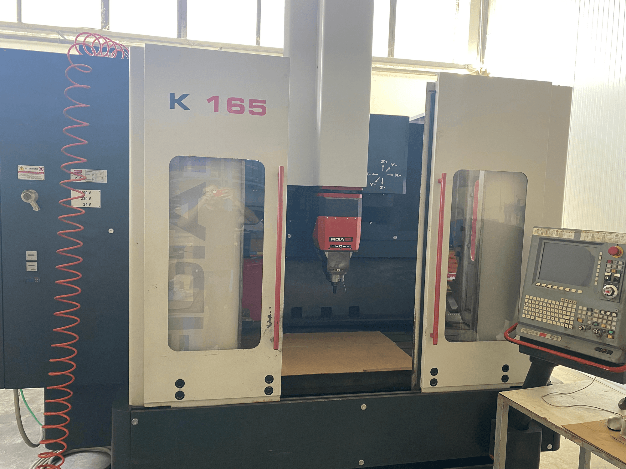 İyi aydınlatılmış bir atölye ortamında önden bakıldığında kırmızı iş mili ve kontrol paneline sahip Fidia K 165 CNC makinesi.