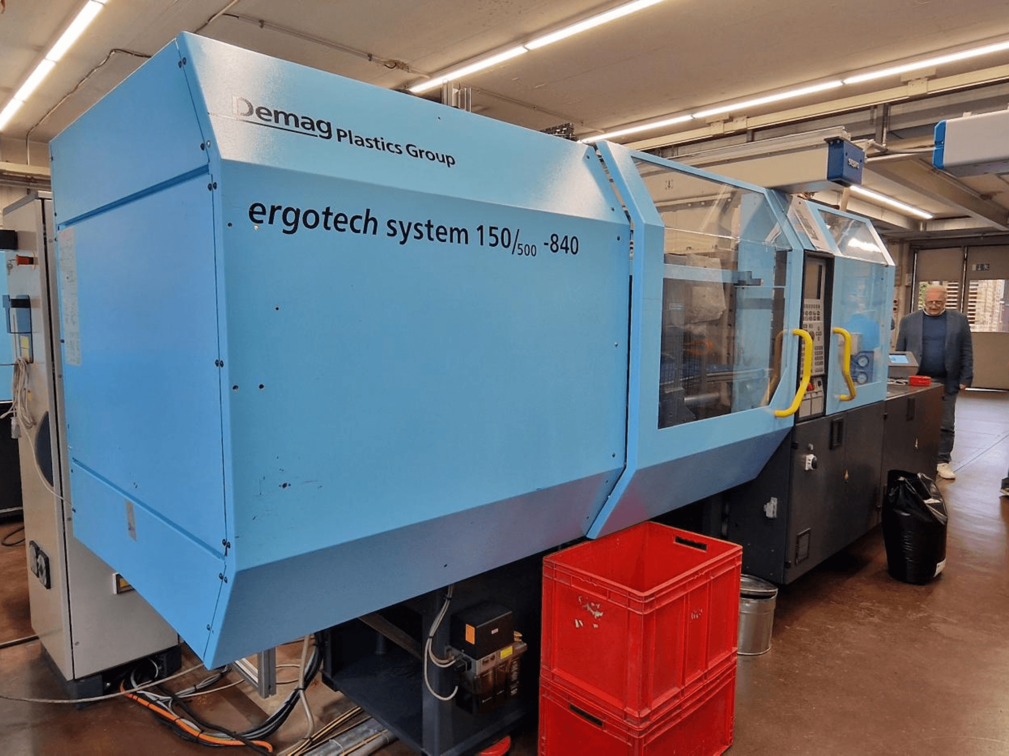 Demag Ergotech System 150/500-840 mavi enjeksiyon kalıplama makinesi, yandan görünüm, arka planda kasalar ve operatör.