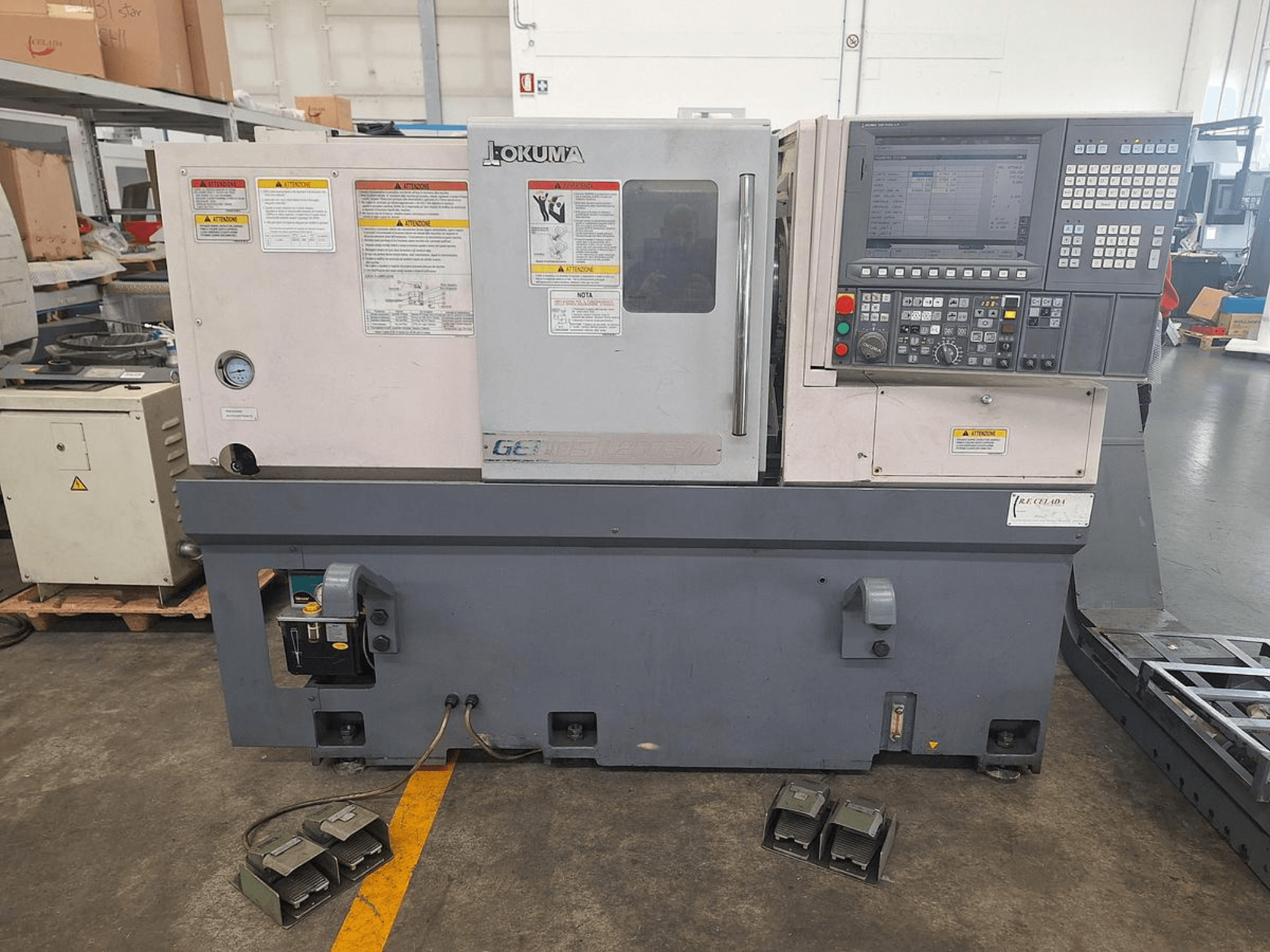 Okuma GENOS L200E-M yatay torna makinesinin yandan görünümü; kontrol paneli, güvenlik uyarıları ve ayak pedallarına sahiptir.
