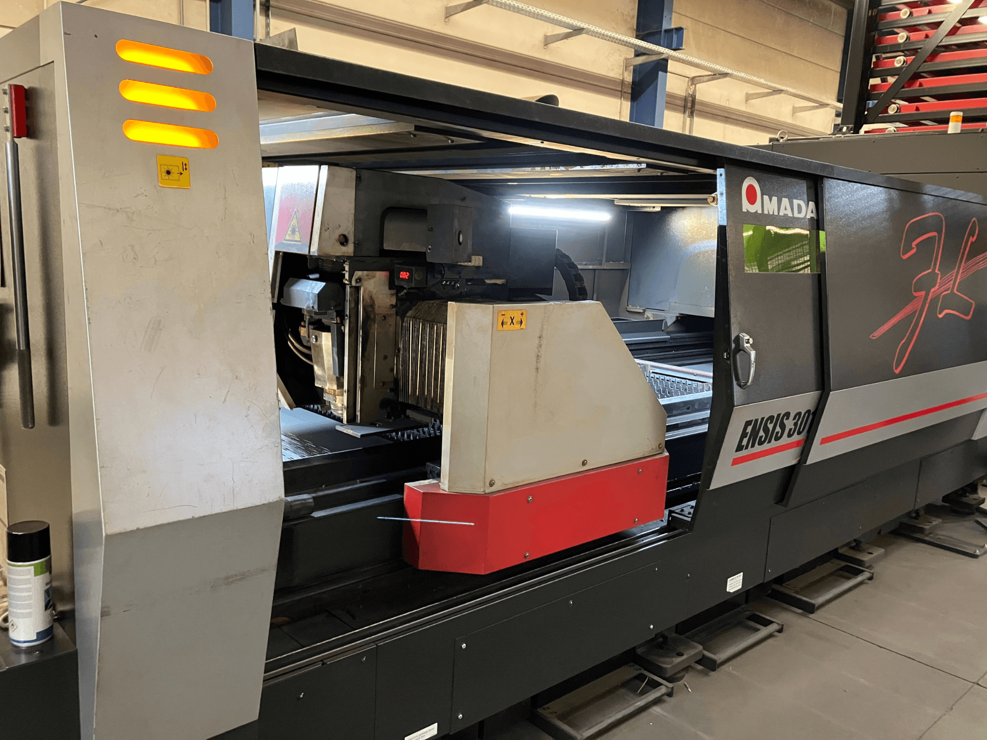 AMADA ENSIS 3015 AJ fiber lazer kesim makinesi, önden görünüm, metal bir çerçeve içinde kesme kafası ve çalıştırma kontrollerini gösterir.