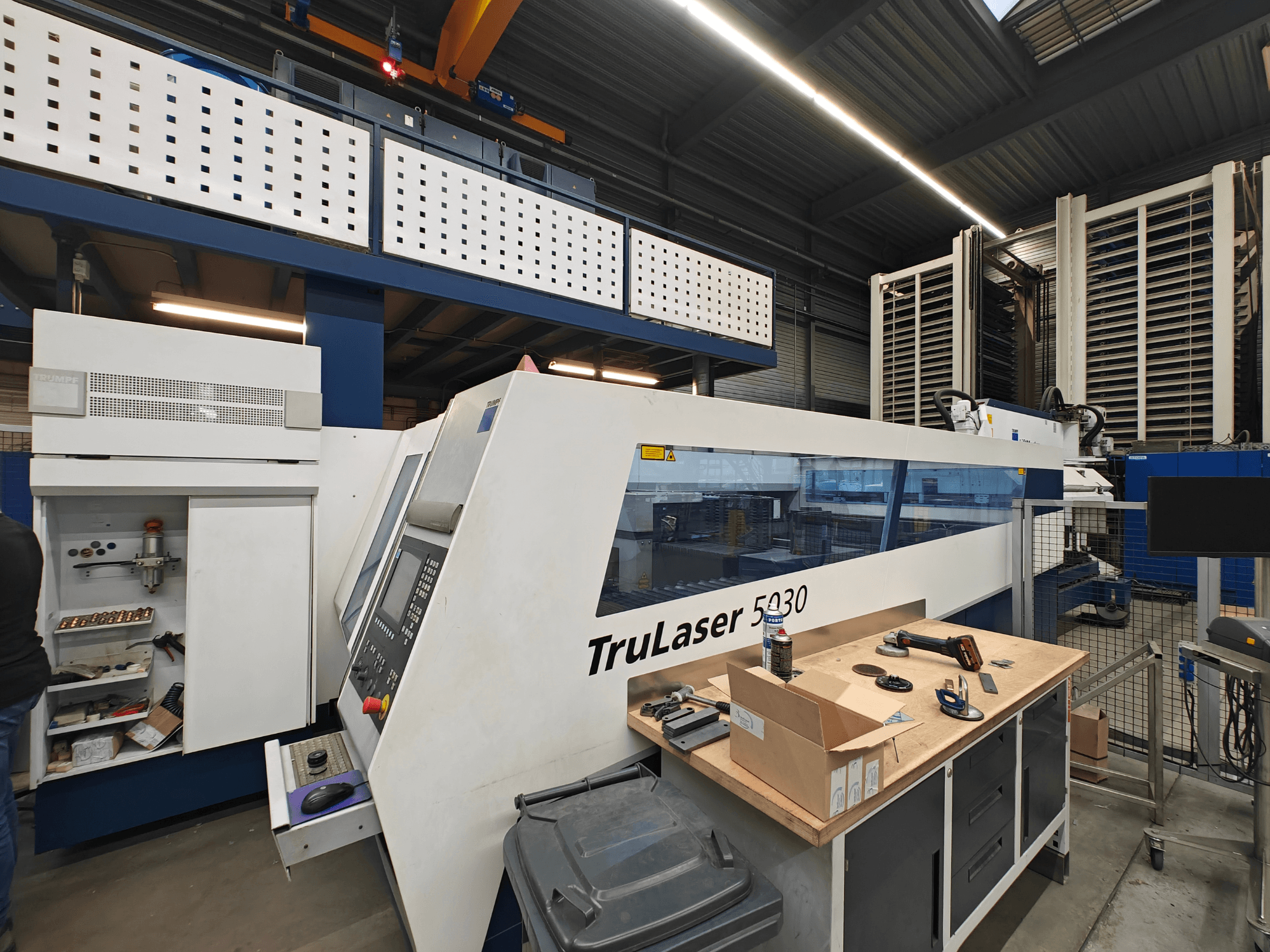 TruLaser 5030 makine bir atölyede, önden görünüm, kontrol paneli görünüyor, etrafı alet ve ekipmanlarla çevrili.