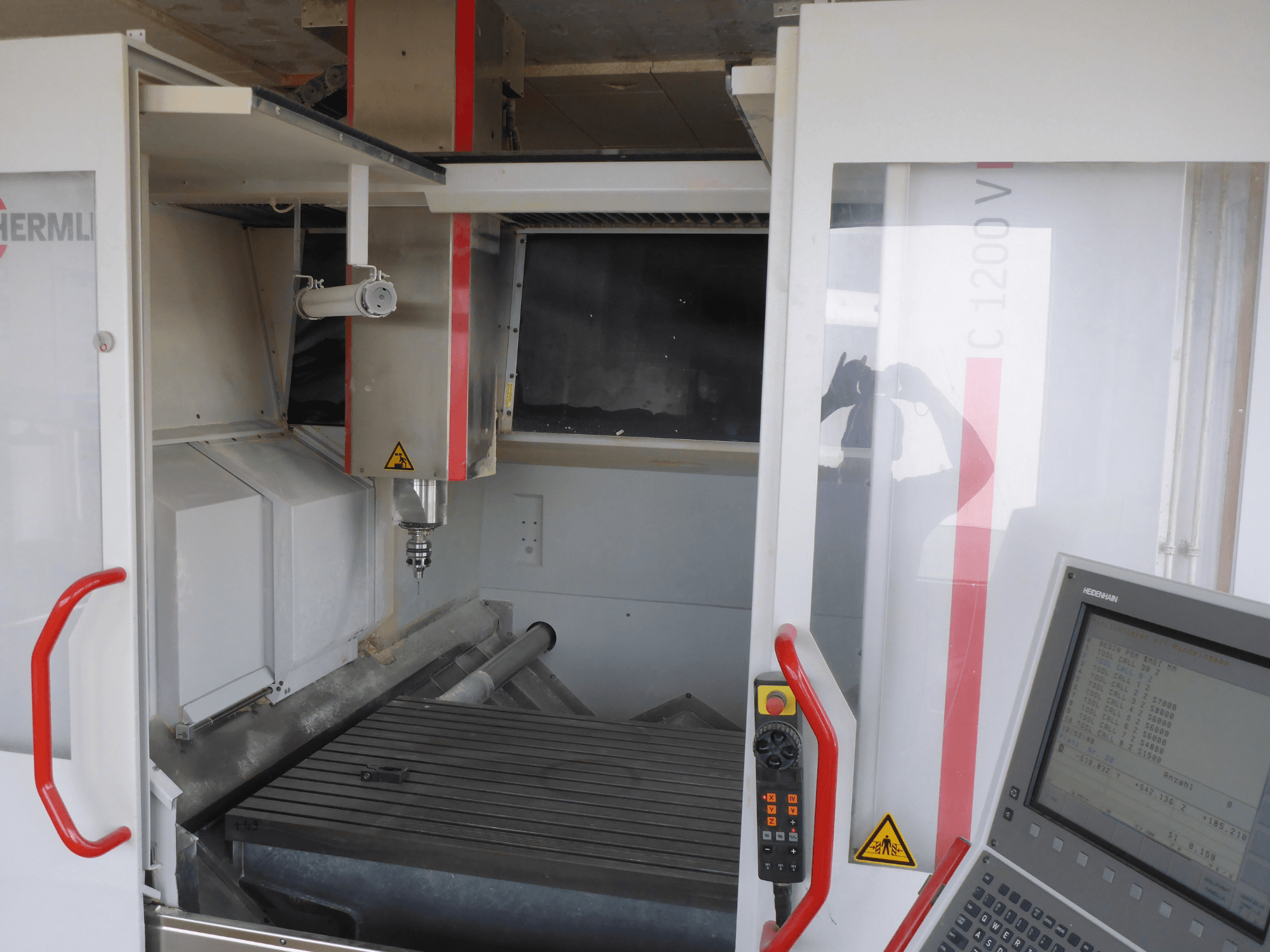 HERMLE C 1200 dikey işleme merkezinin iç kısmında takım tutucu, kontrol paneli ve çalışma tablasının önden görünümü.