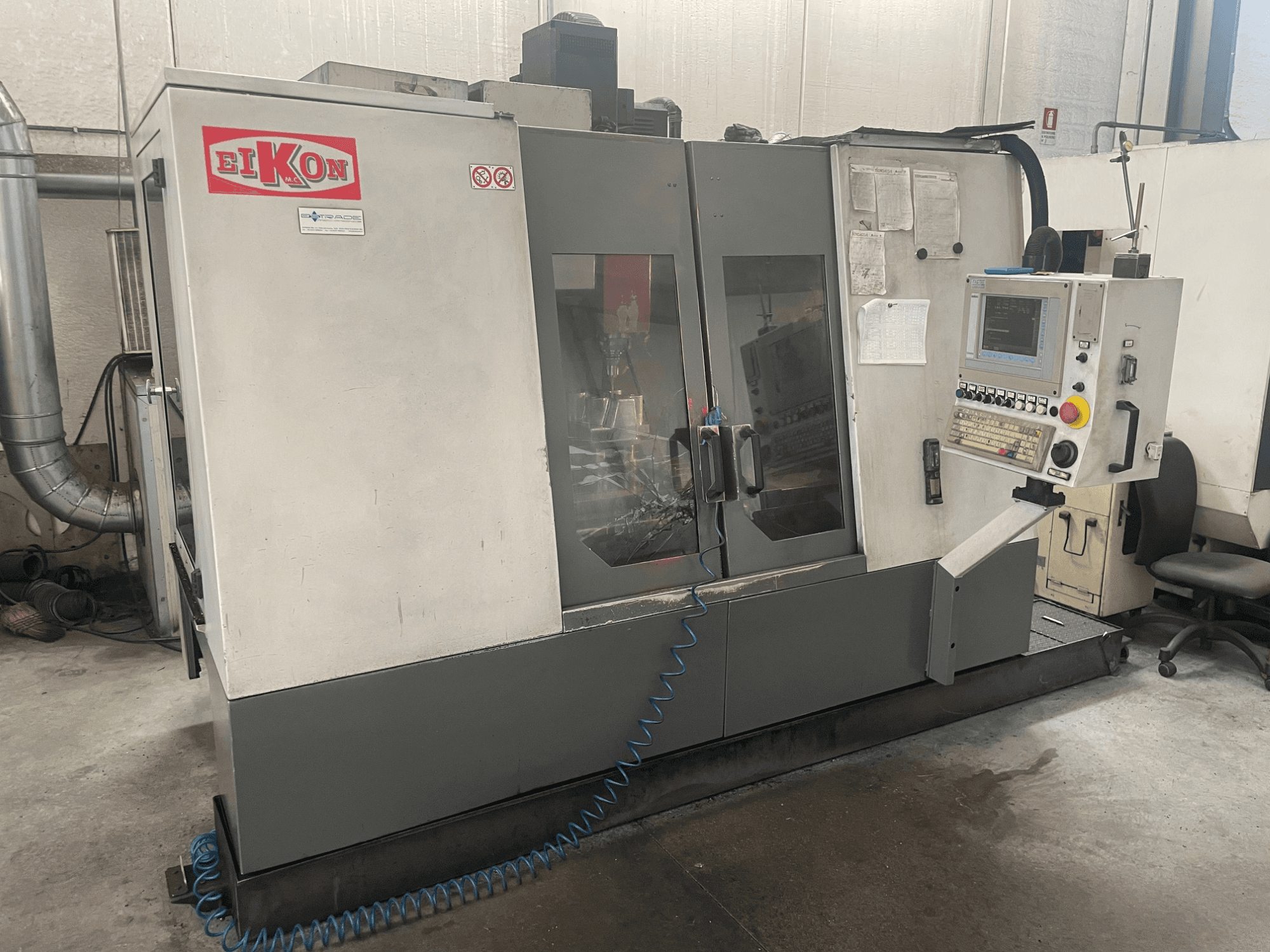 Kontrol paneli, büyük cam kapılar ve zeminde mavi bir hava hortumu bulunan EIKON CNC makinesi önden görüntülenmiştir.