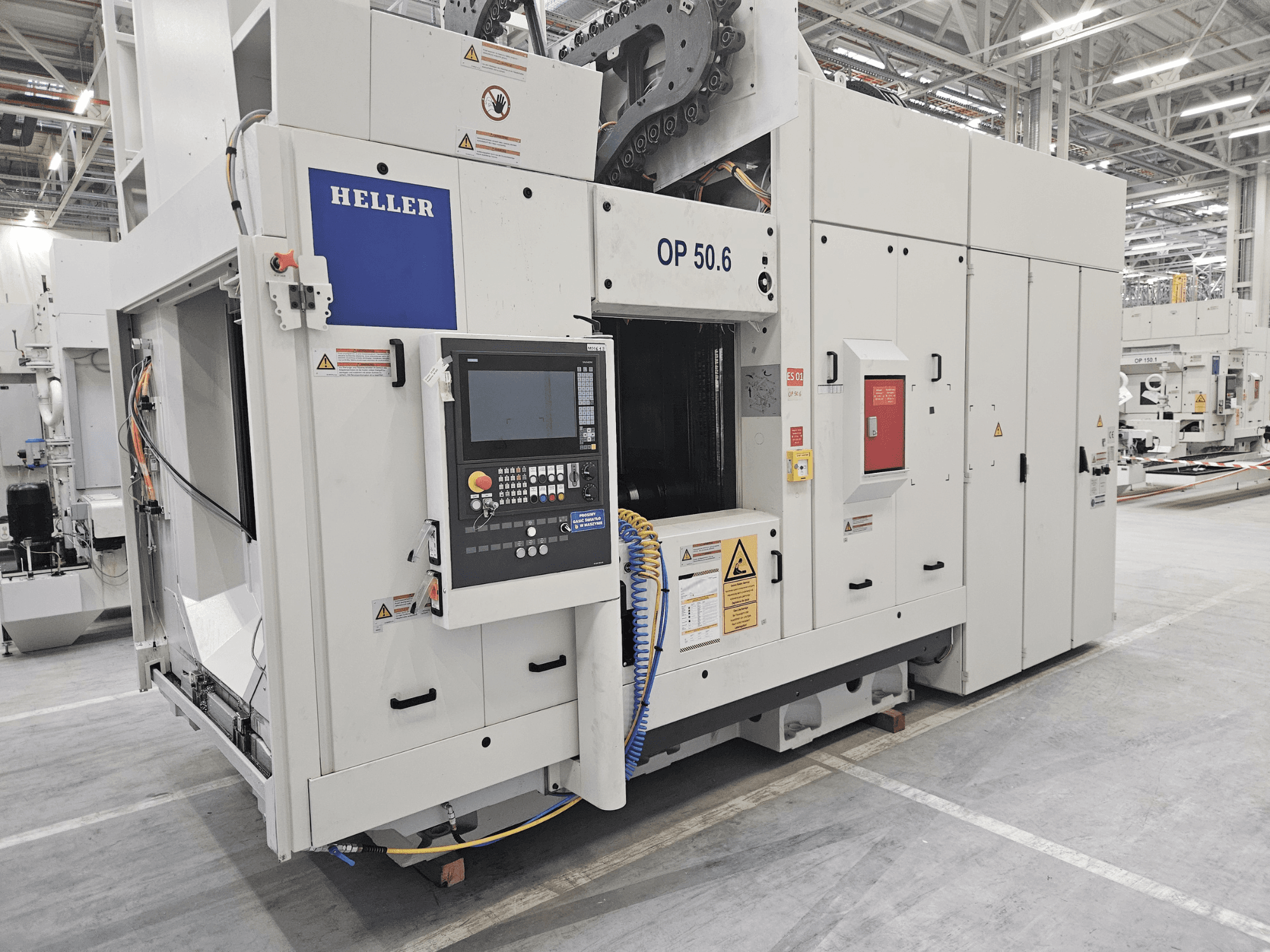 Heller OP 50.6 işleme merkezi, önden görünüm, kontrol paneli, uyarı etiketleri ve hortumlar fabrika ortamında görülebilir.