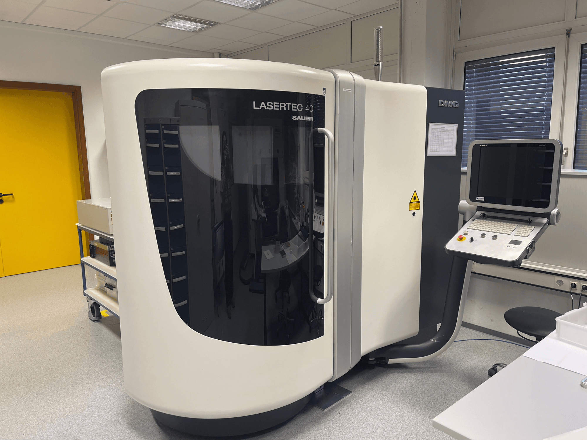 DMG MORI'nin beyaz LASERTEC 40 freze makinesi, önden görünüm, iyi aydınlatılmış bir odada kontrol paneli ve ergonomik tasarıma sahiptir.