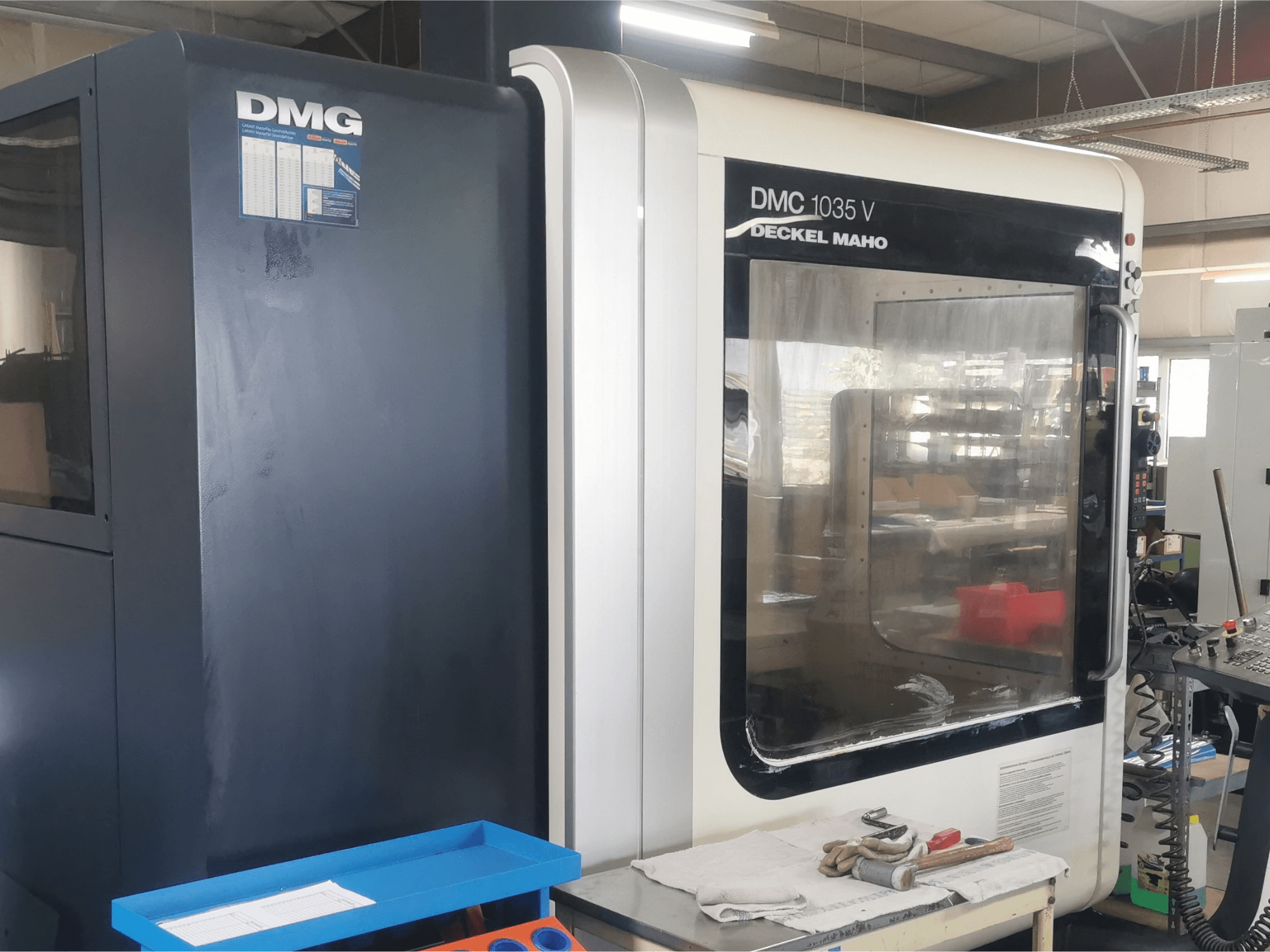 Atölyede DMG DMC 1035 V freze makinesi, mavi takım tepsisi ve yüzeyde eldivenlerle önden görünüm.
