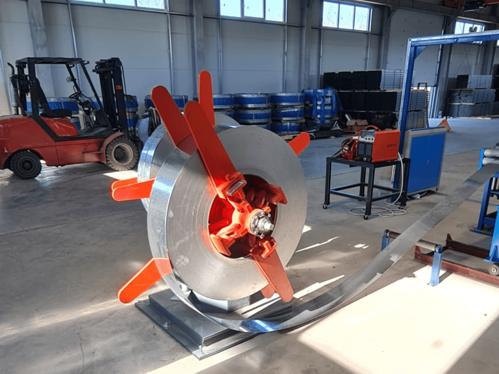 Bir stand üzerinde kırmızı saplı metal bobin, bir açıdan görüntüleniyor. Makineler ve bir forklift ile endüstriyel arka plan.