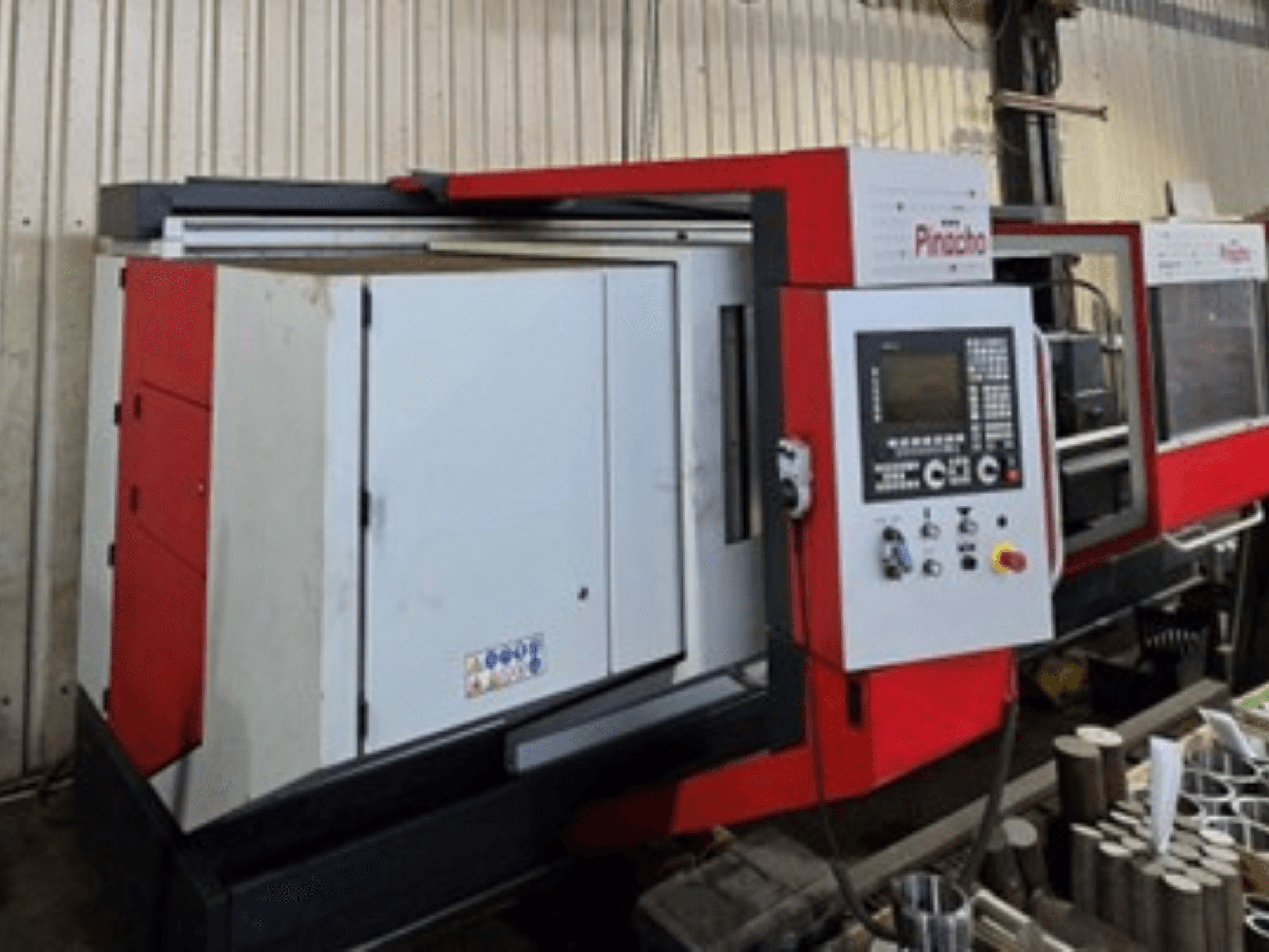 Kırmızı ve beyaz Pinacho CNC makinesi, yandan görünüm, kontrol paneli ve zemindeki malzemeler ile çalışma kurulumunu gösterir.