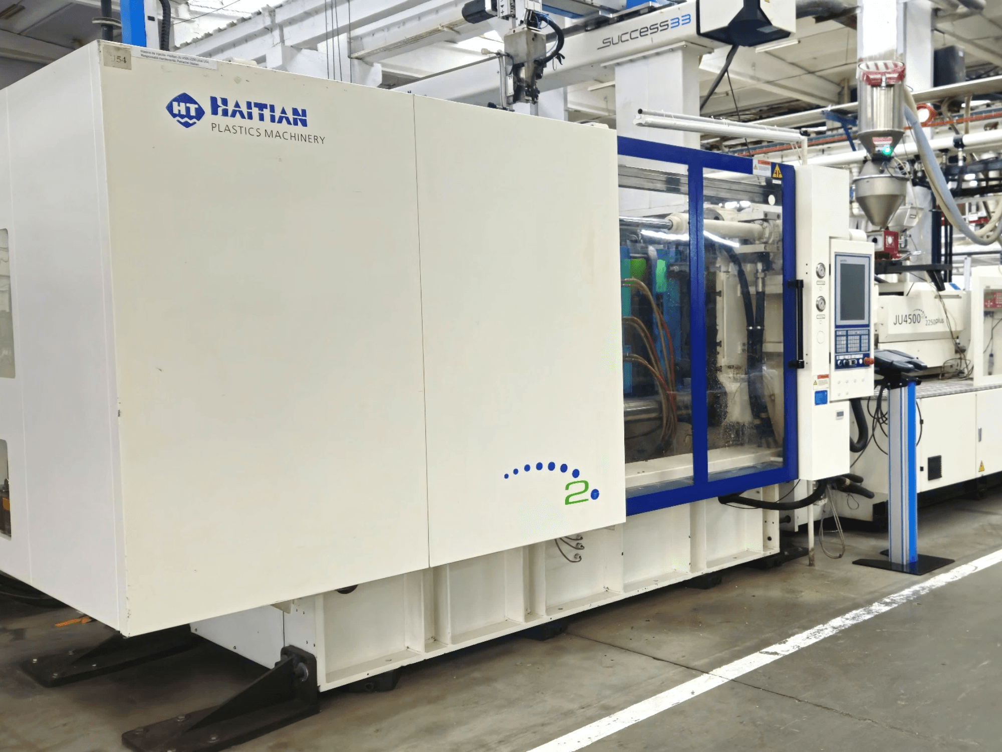 Haiti Plastik Makineleri modeli, önden görünüm, kontrol panelli ve görünür hortumlu makineleri endüstriyel bir ortamda sergiliyor.