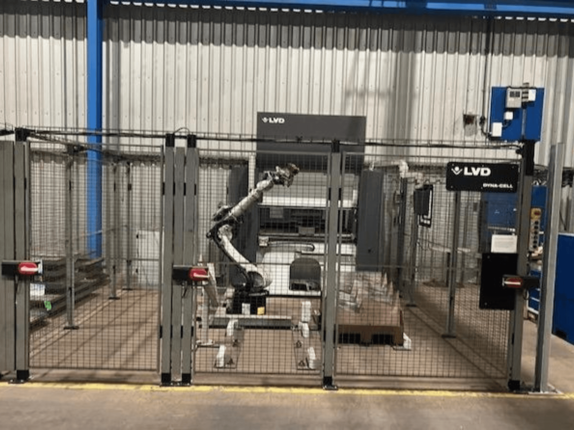 Çitle çevrili bir muhafaza içindeki LVD Dyna-Cell robotik iş istasyonu, önden görünüm, robotik kol ve kontrol paneli.